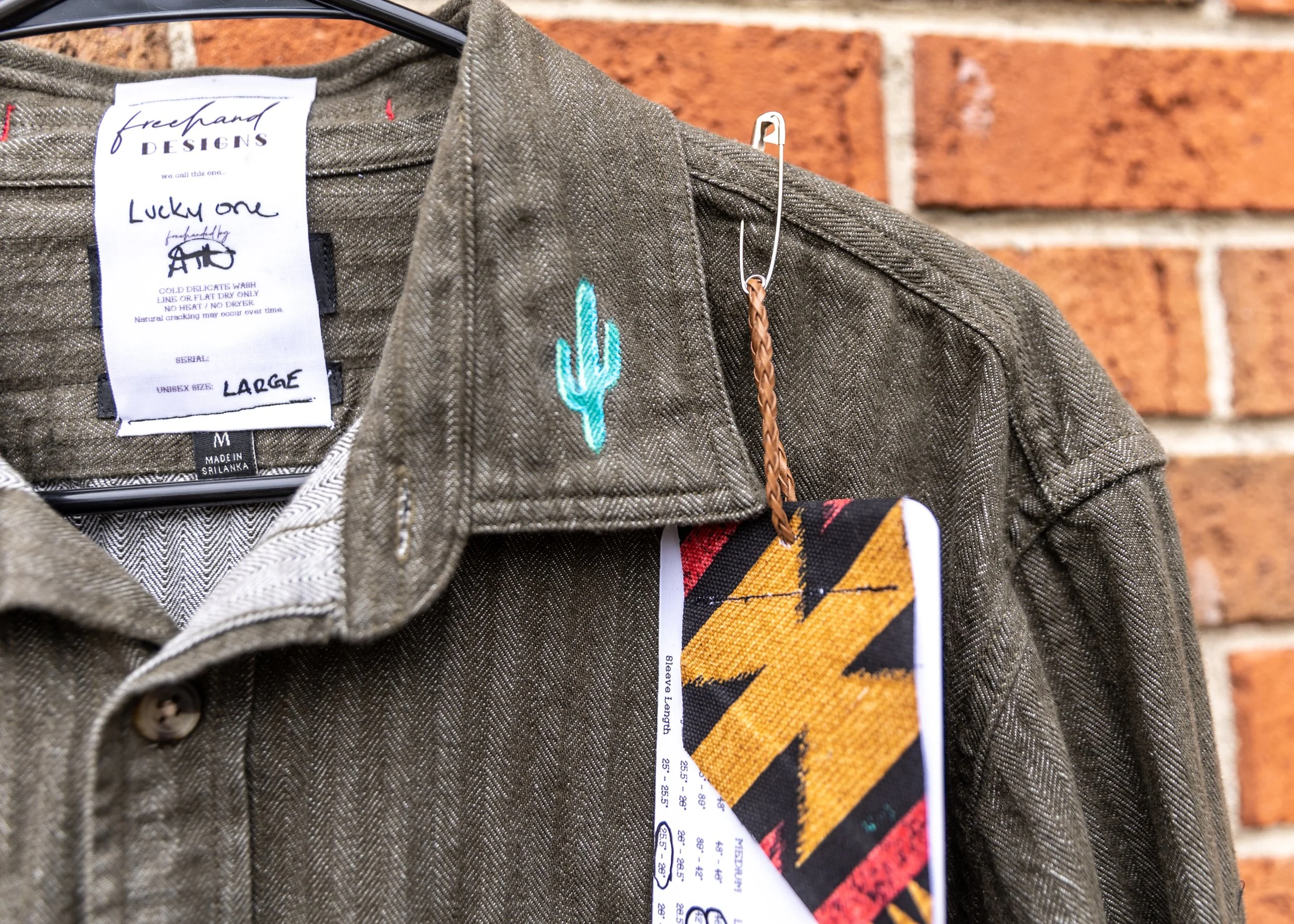 Close-up of a clothing tag on a brown jacket with a light blue cactus embroidered on the shoulder area, against a brick wall background.