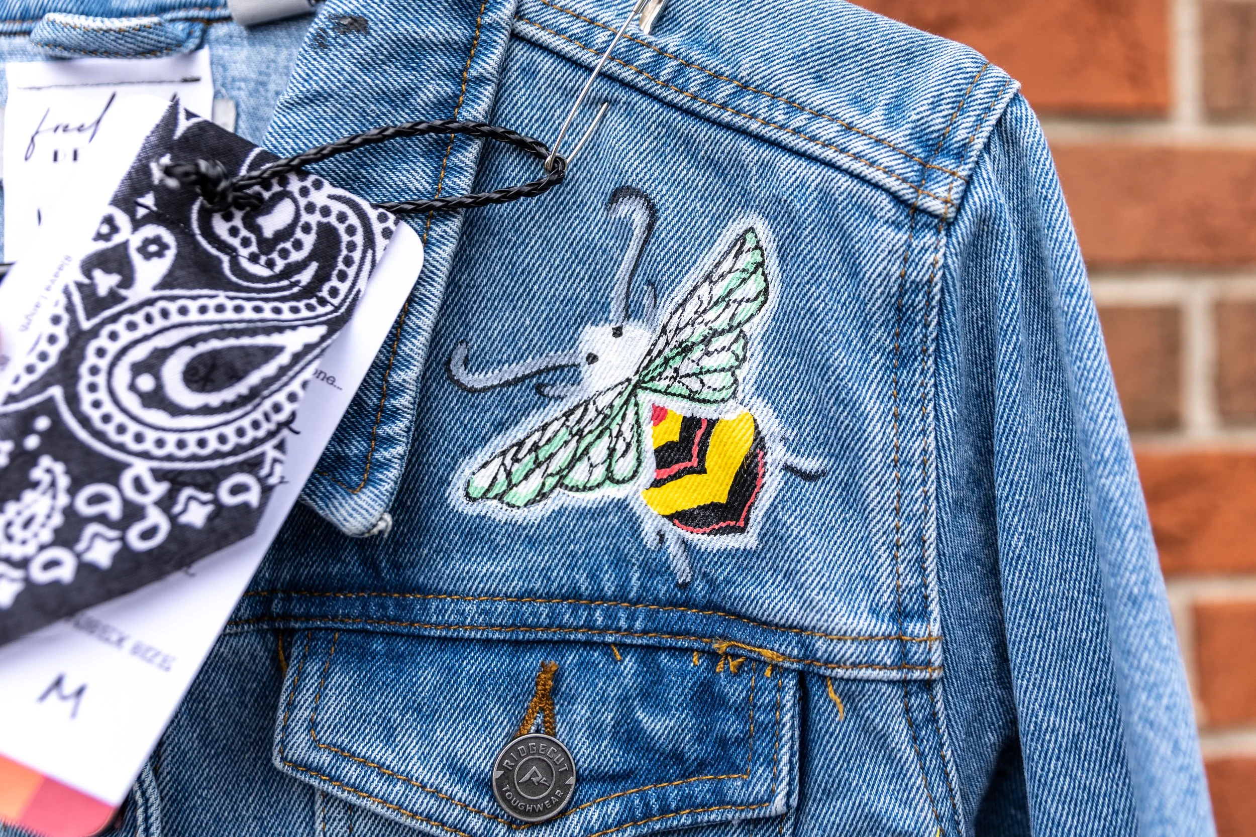 Close-up of a denim jacket with an embroidered butterfly patch, a tag attached with a black bandana-style string, and a metal button.