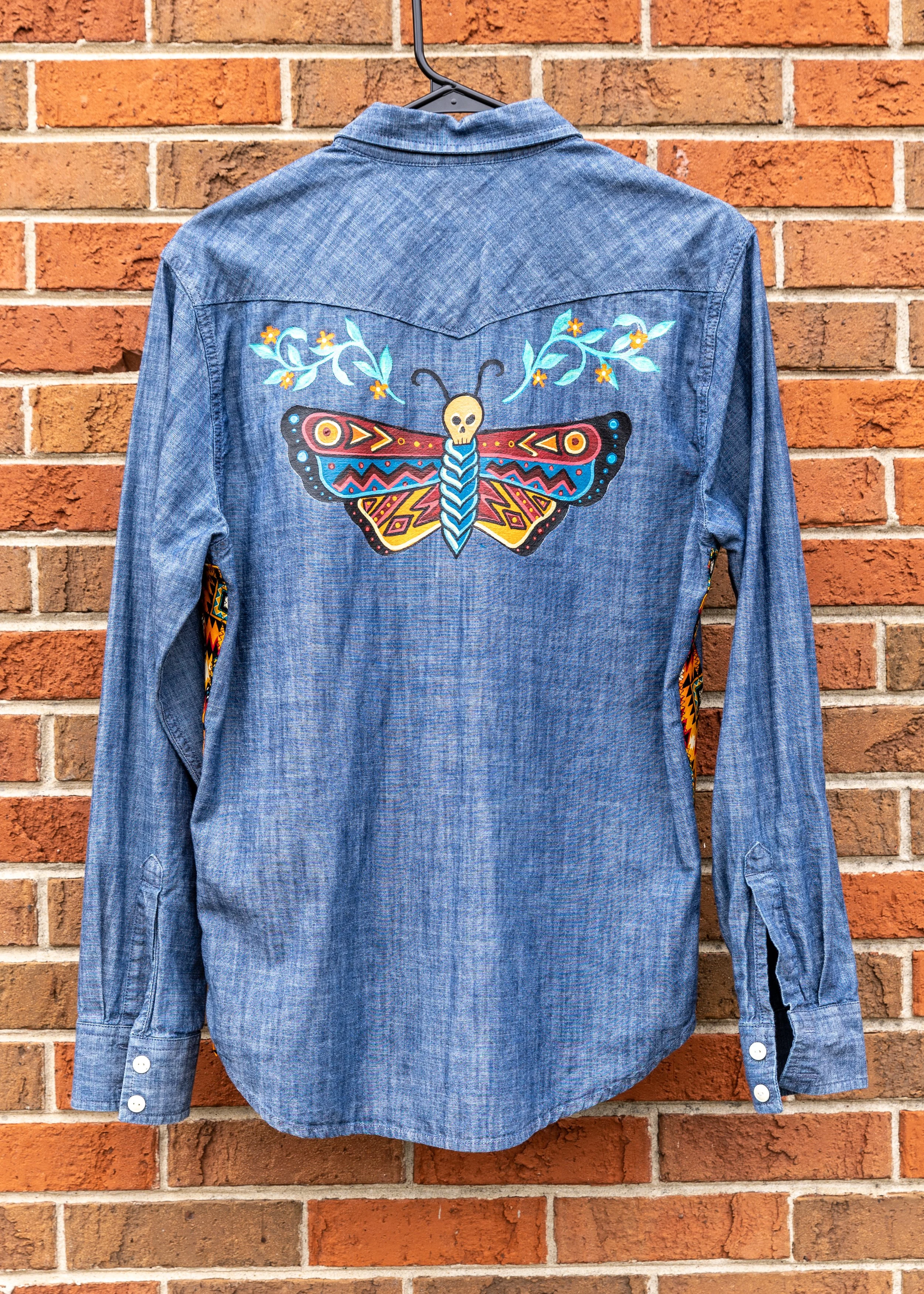 Back of a denim shirt with colorful embroidered butterfly and floral design on the upper back, hanging on a hanger against a brick wall.
