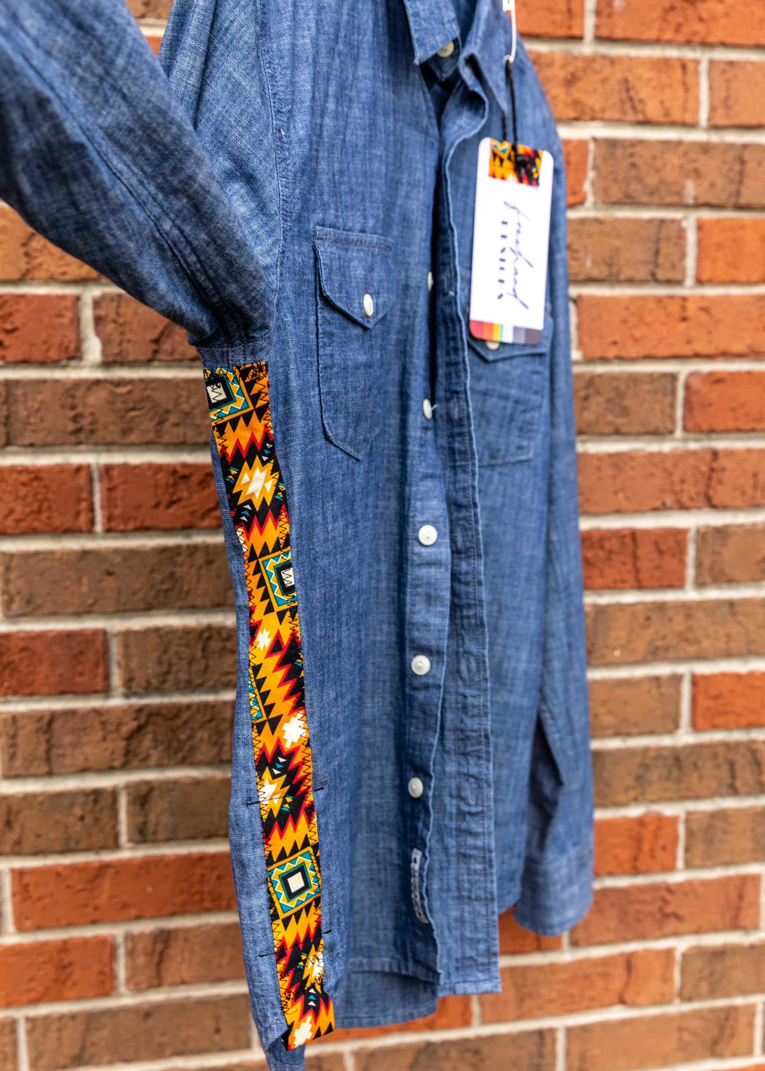 Close-up of a blue denim shirt with a vibrant, multicolored tribal pattern on the sleeve seam, worn by a person standing against a red brick wall.
