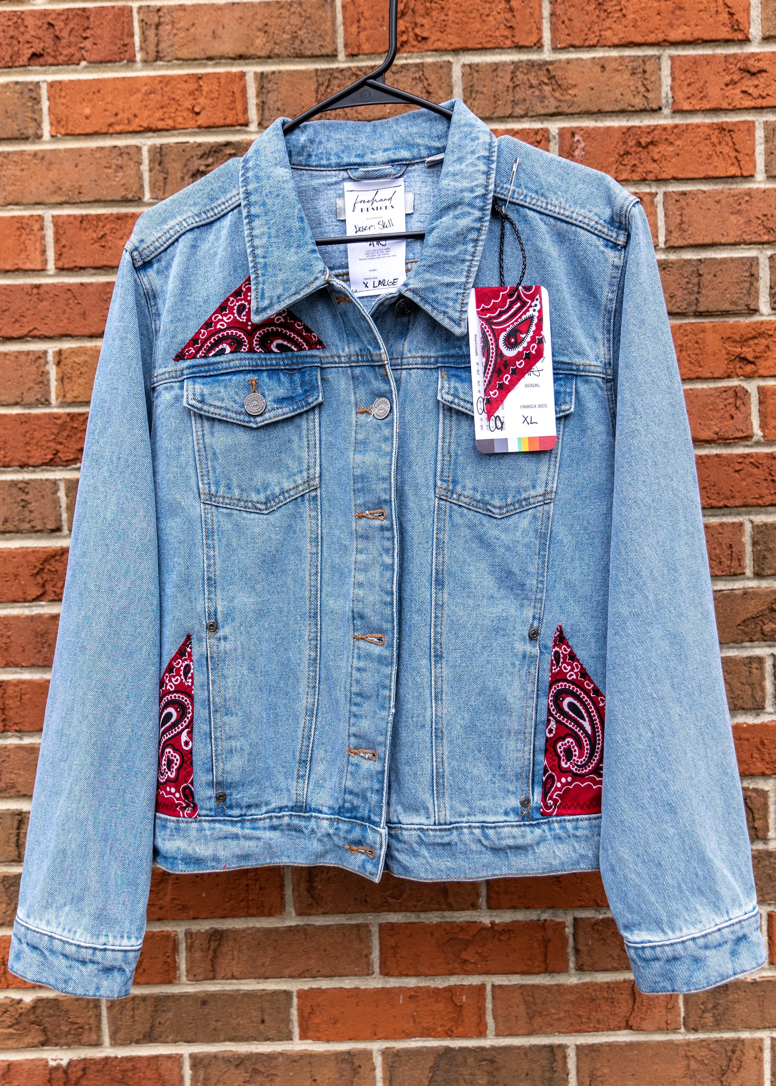 A light blue denim jacket with red paisley-patterned fabric accents on the shoulders and sides, hanging on a black hanger against a brick wall.