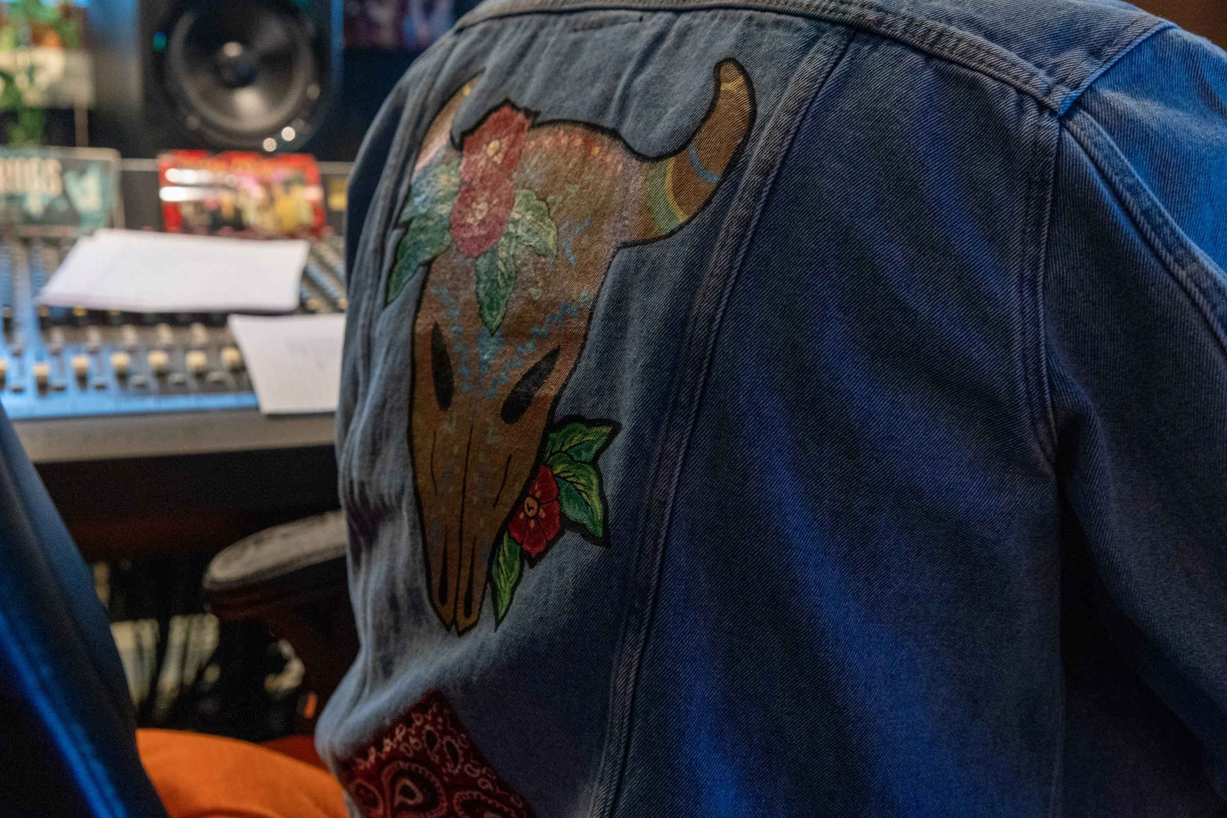 Close-up of a denim jacket with a colorful embroidered patch of a dog's face on the shoulder, with blurred background including audio equipment and papers.