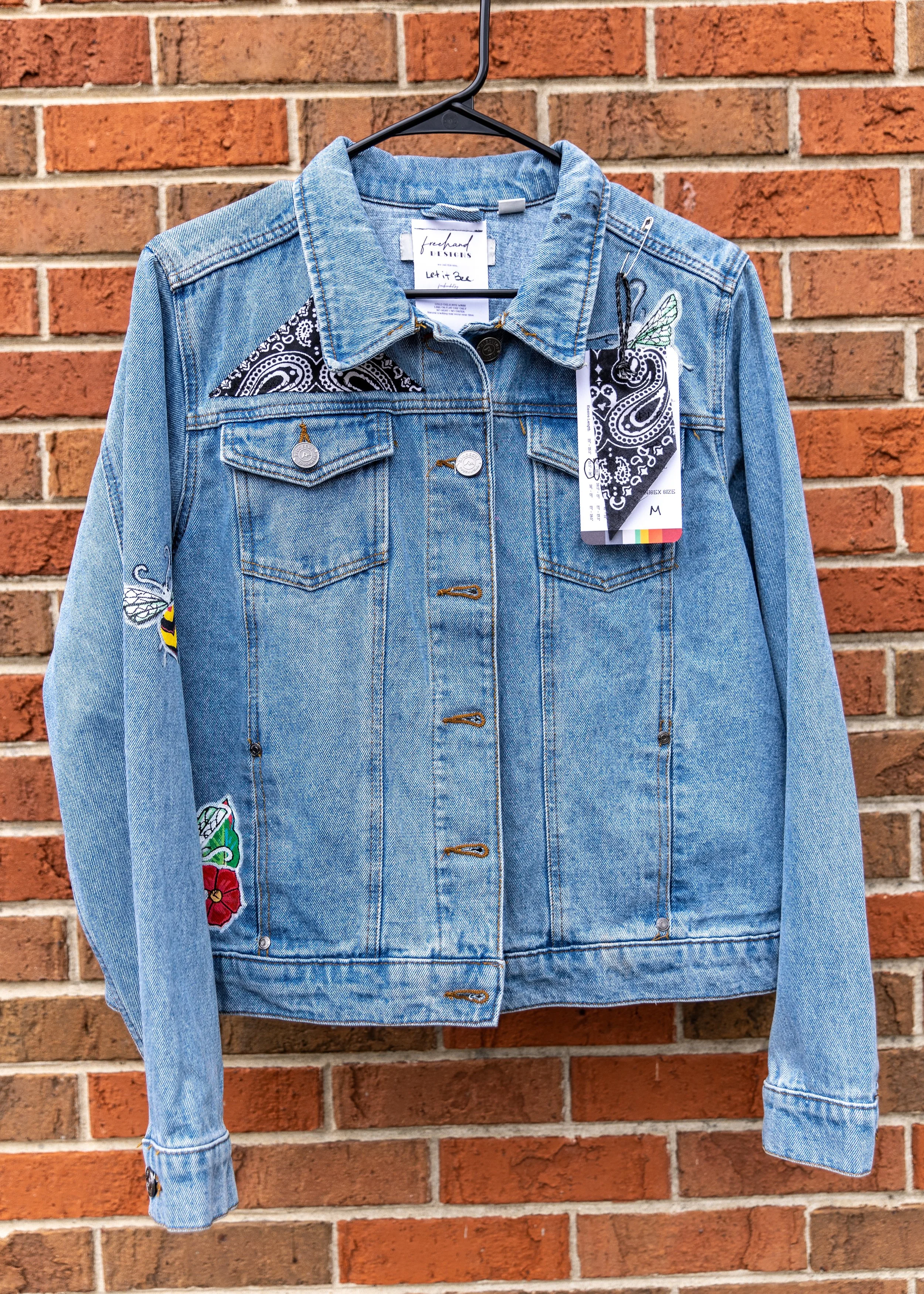 Light blue denim jacket with embroidered patches on the front, hanging on a black hanger against a red brick wall.