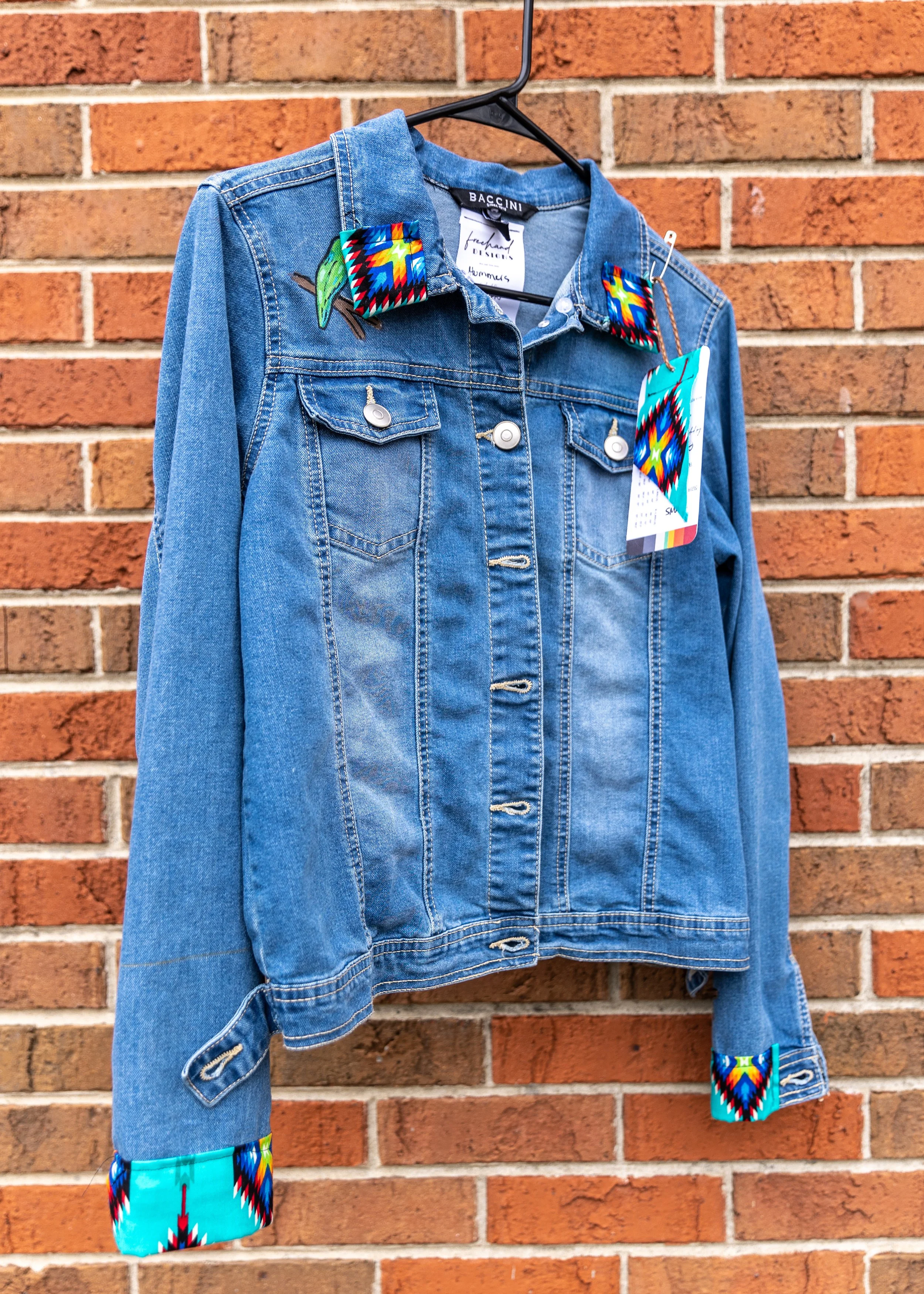 Blue denim jacket hanging on a black hanger against a red brick wall, with colorful Southwestern-patterned fabric accents on the collar, cuffs, and a tag.
