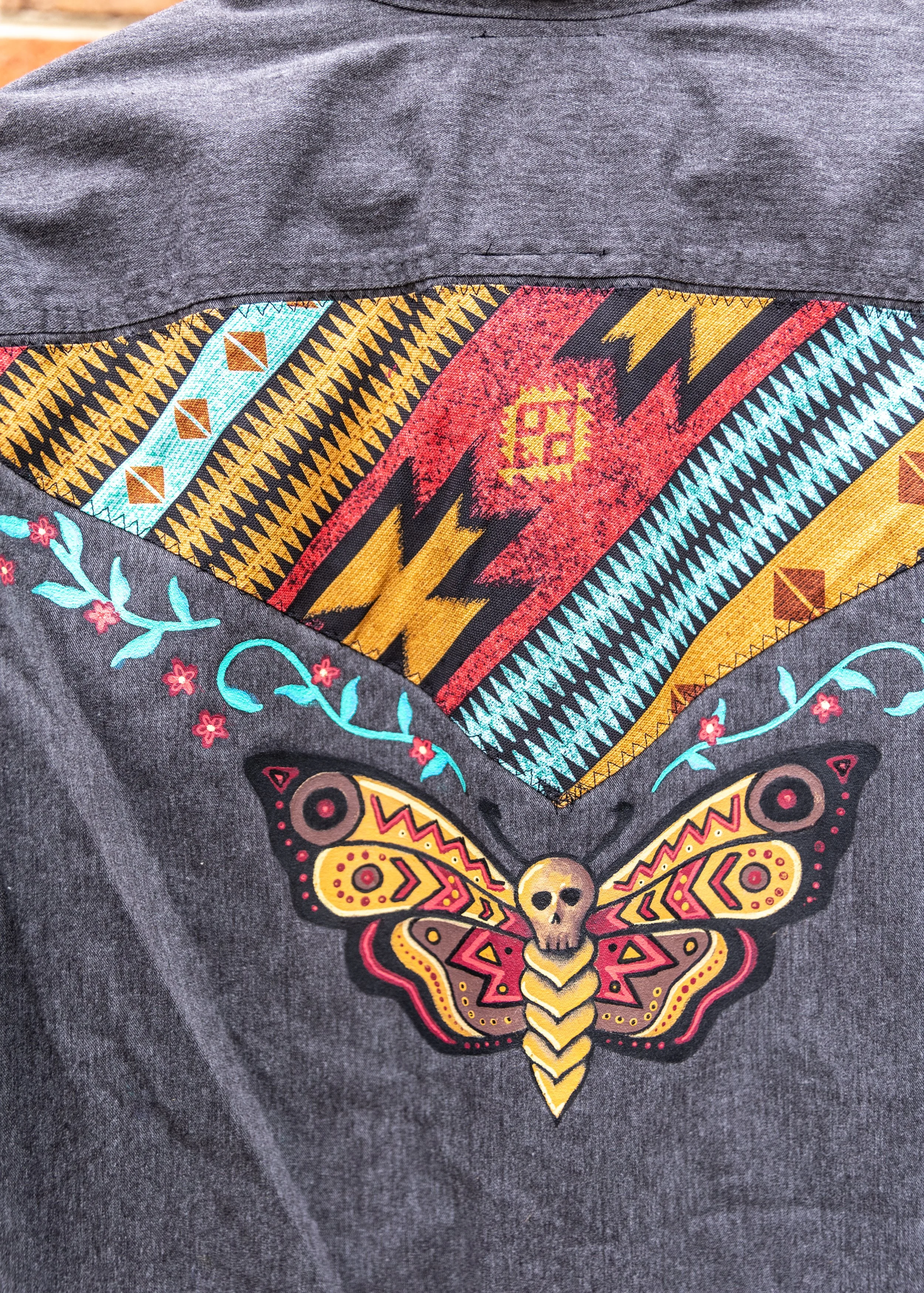 A dark gray fabric with a colorful embroidered design of a butterfly with a skull at its center, surrounded by floral and geometric patterns.