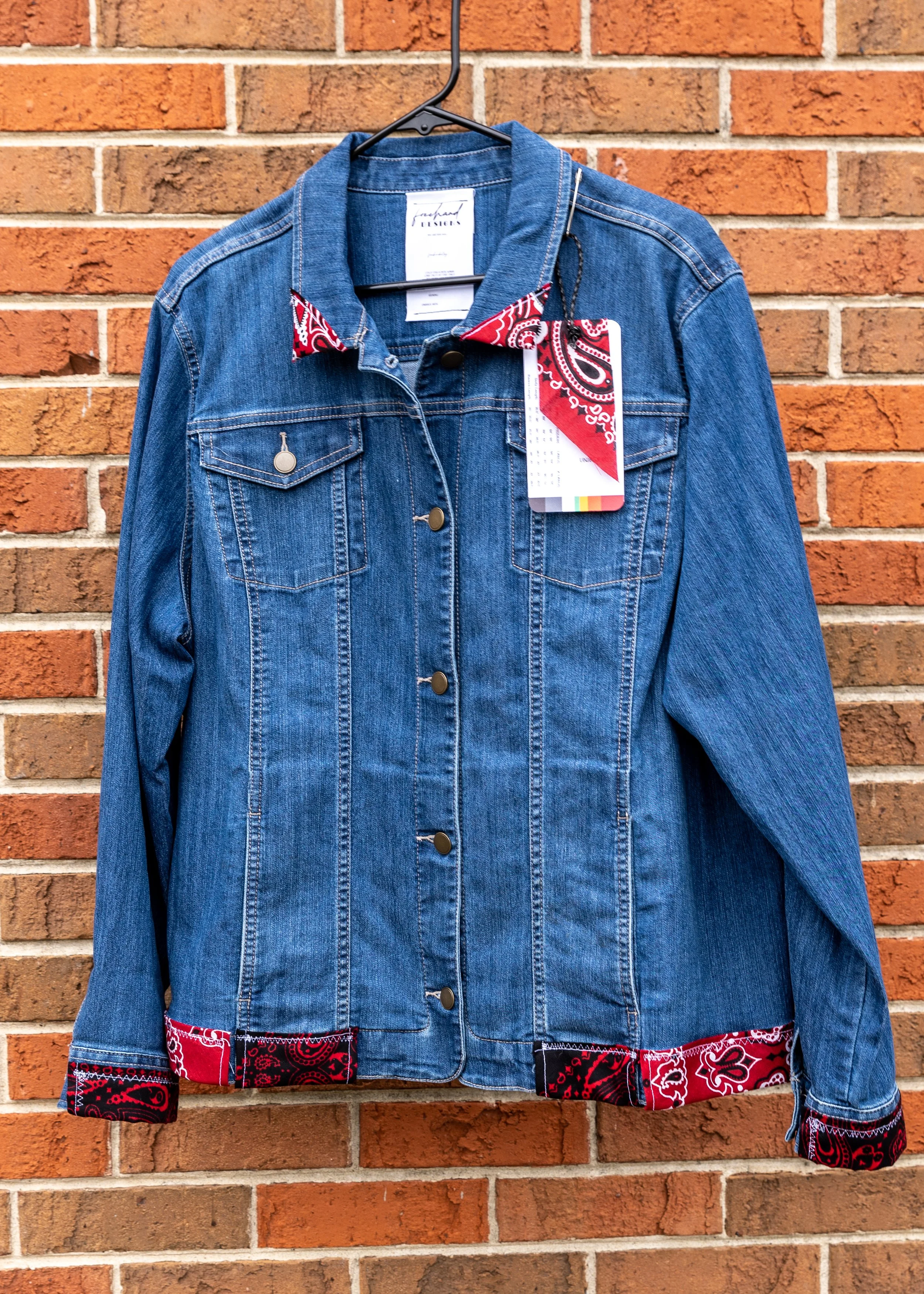 A denim jacket with red bandana fabric accents on the collar, cuffs, and bottom hem, hanging on a hanger against a brick wall.