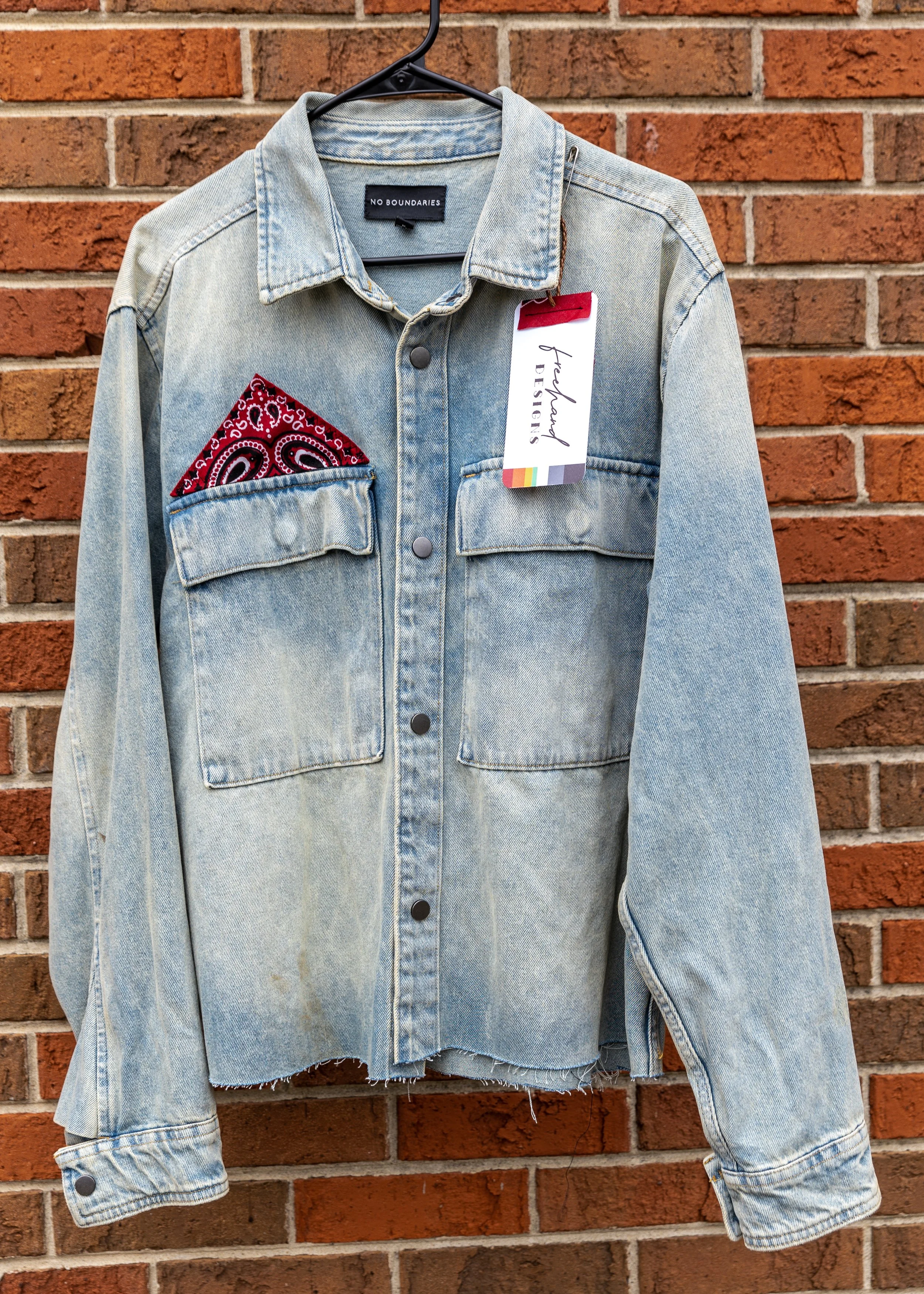 Light blue denim jacket hanging on a black hanger against a brick wall. The jacket features a red bandana in the left chest pocket, a tag attached to the collar with the text 'hand DESIGNS', and a black label inside the collar with the text 'NO BOUND