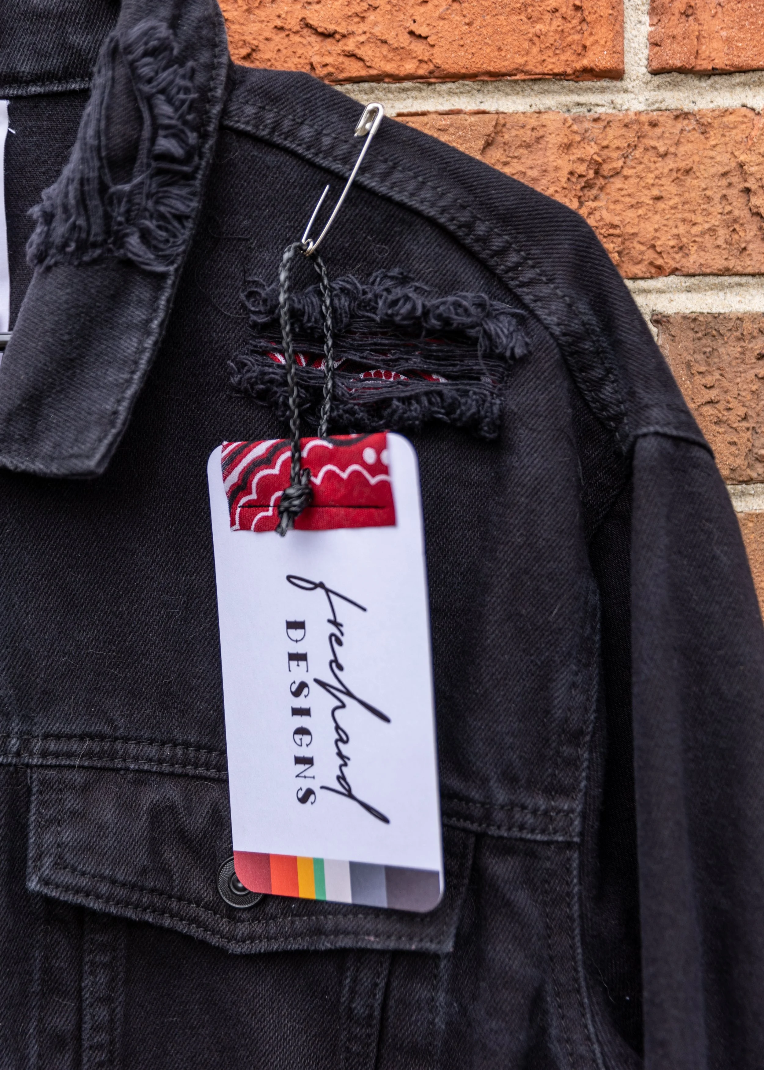 Close-up of a black denim jacket with a colorful tag attached, hanging against a brick wall.