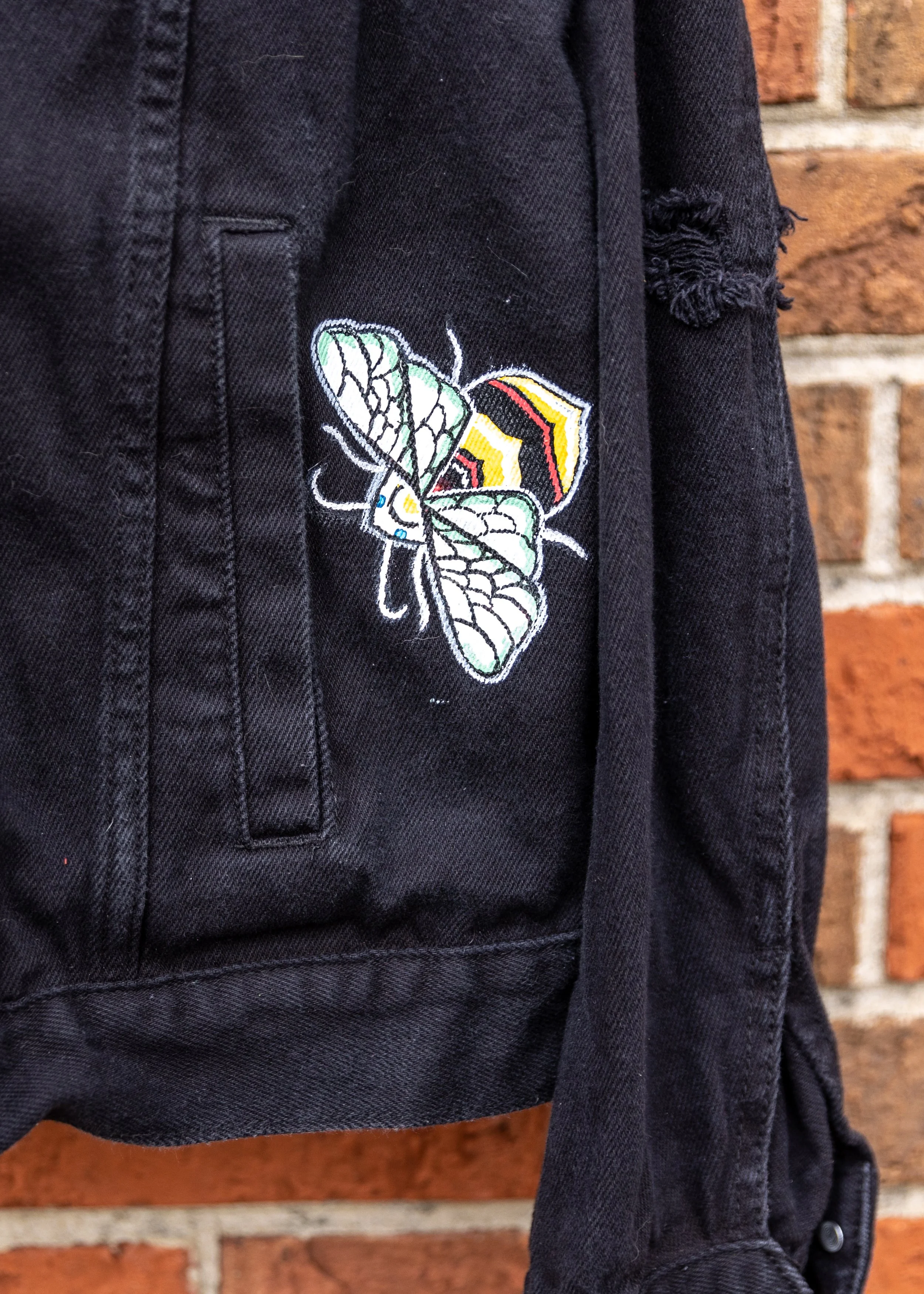 Close-up of a black denim jacket with a butterfly embroidered design on the pocket, set against a brick wall background.