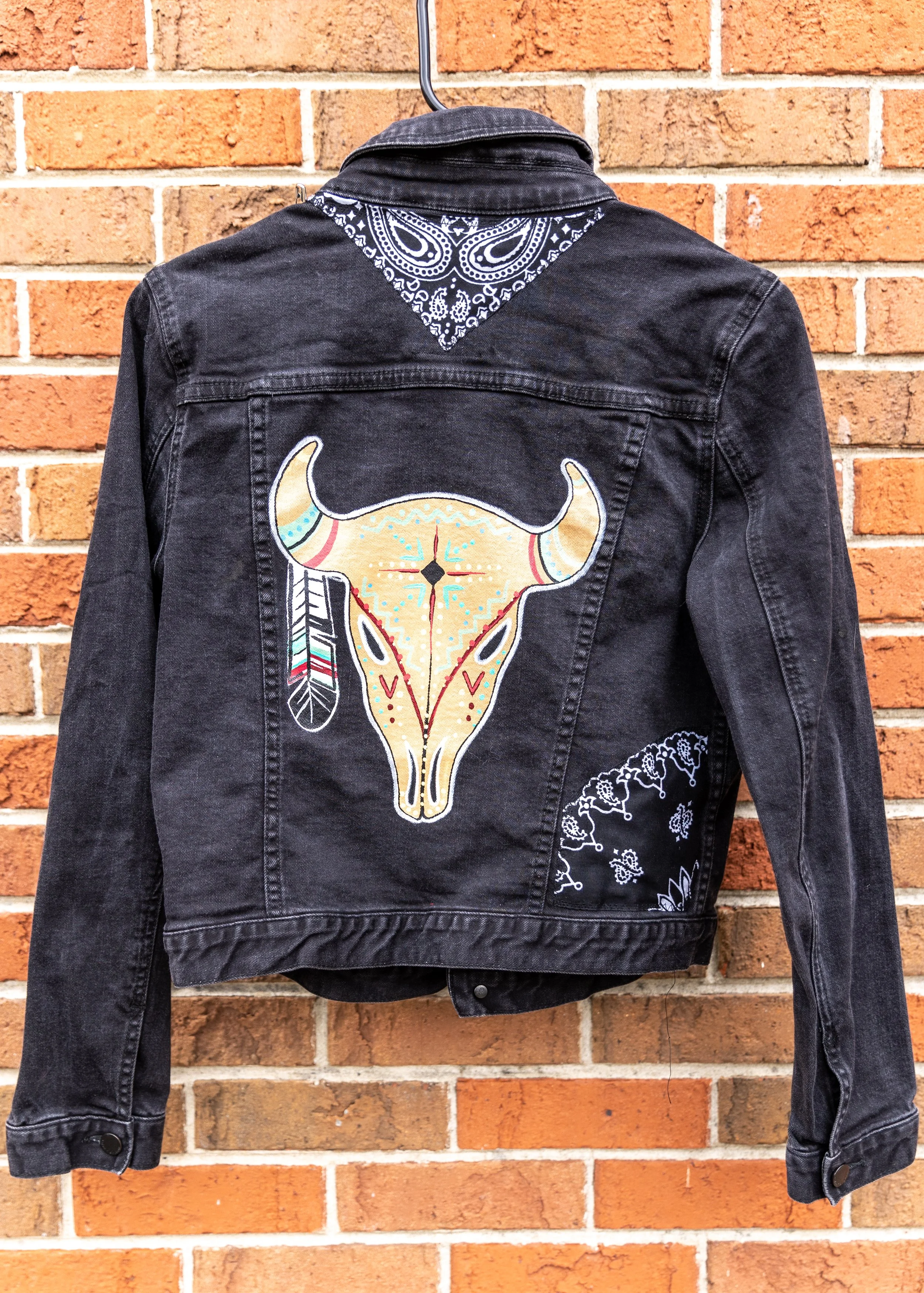 Black denim jacket with a large painted buffalo skull design on the back, topped with a blue bandana with white paisley pattern, hanging against a brick wall.
