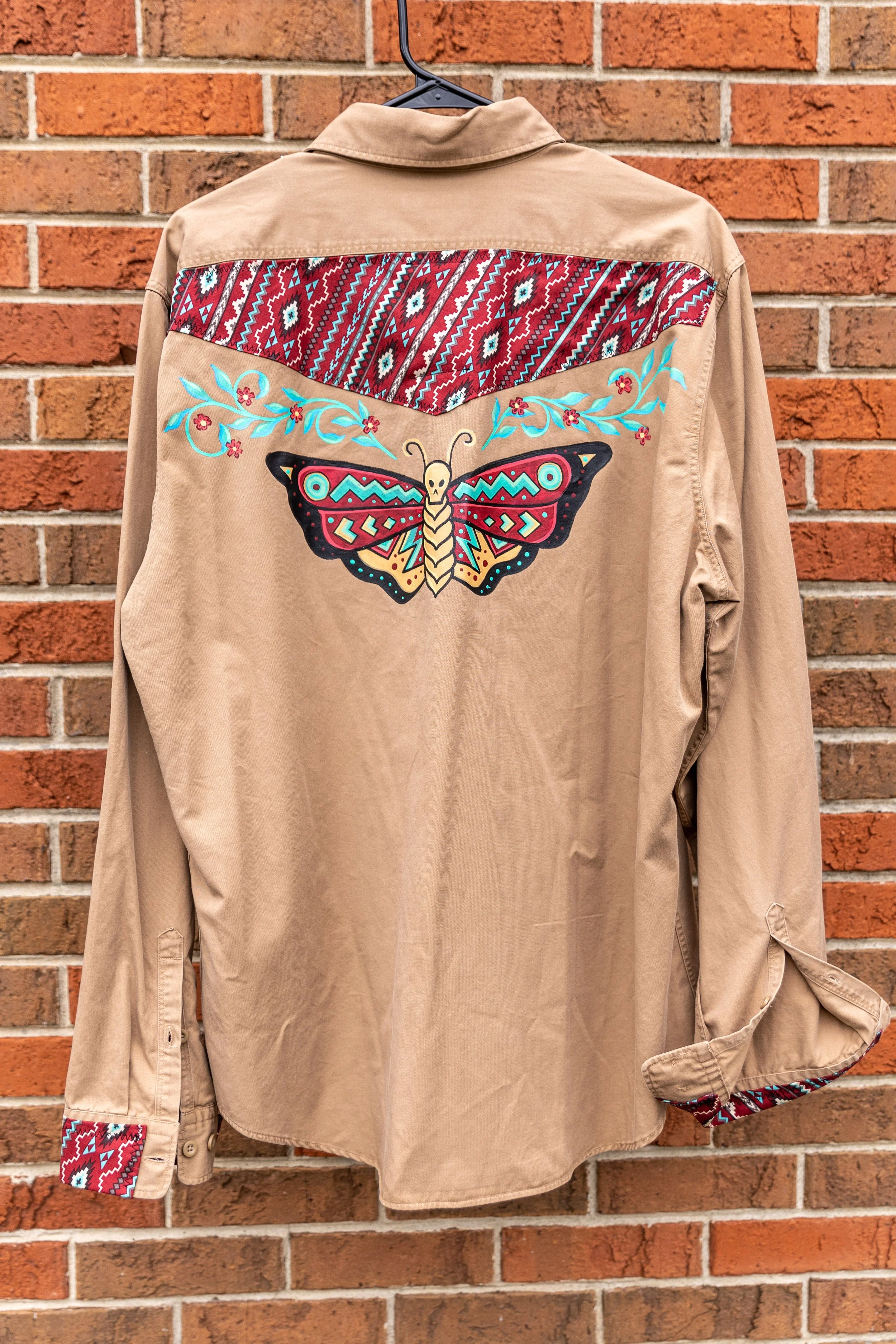 Beige shirt with colorful embroidered butterfly and geometric patterns, hanging on a hanger against a brick wall.