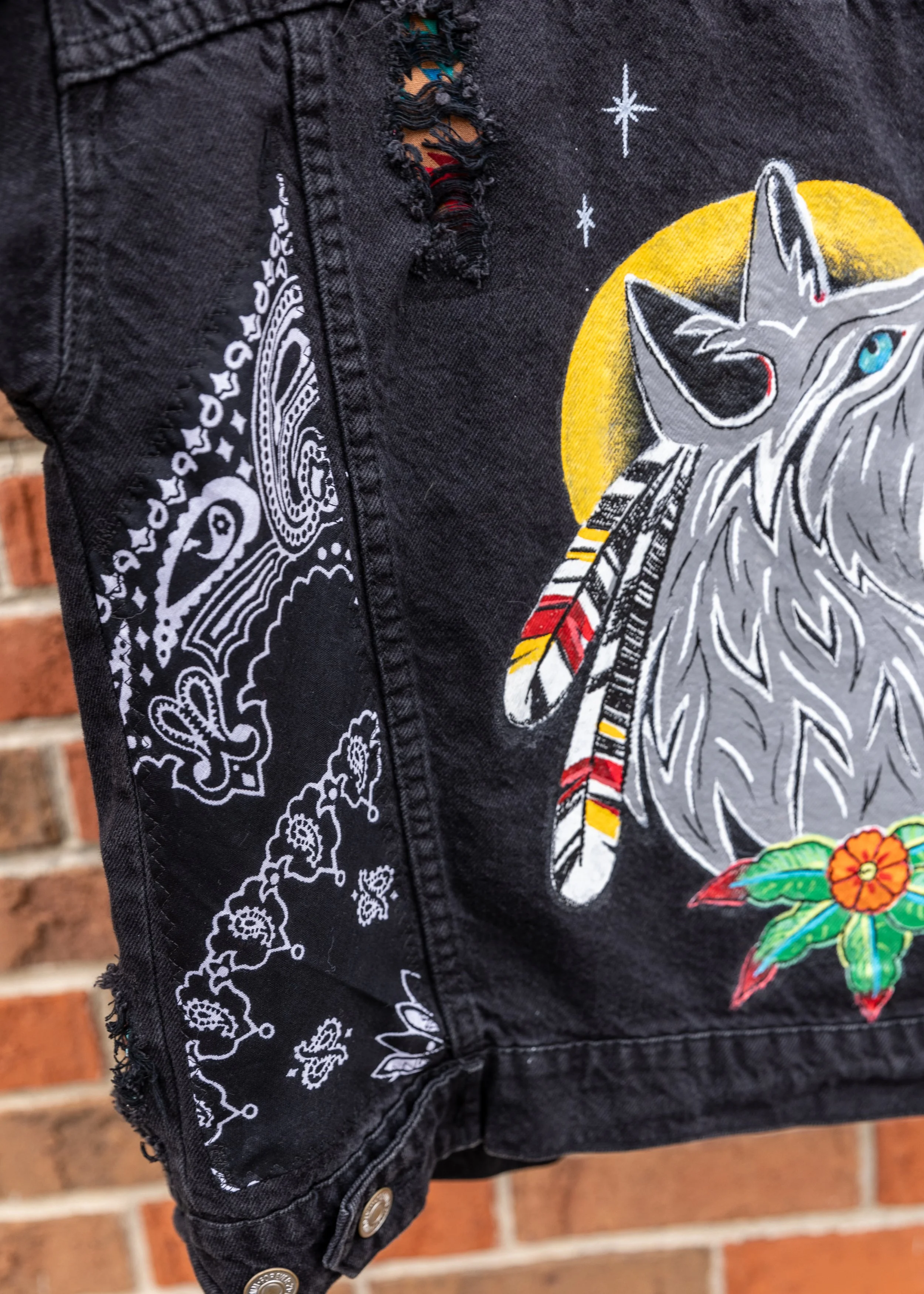Close-up of black denim jacket with embroidered wolf head, moon, stars, and floral designs, featuring a distressed hole near the neckline and silver buttons on the sleeve.