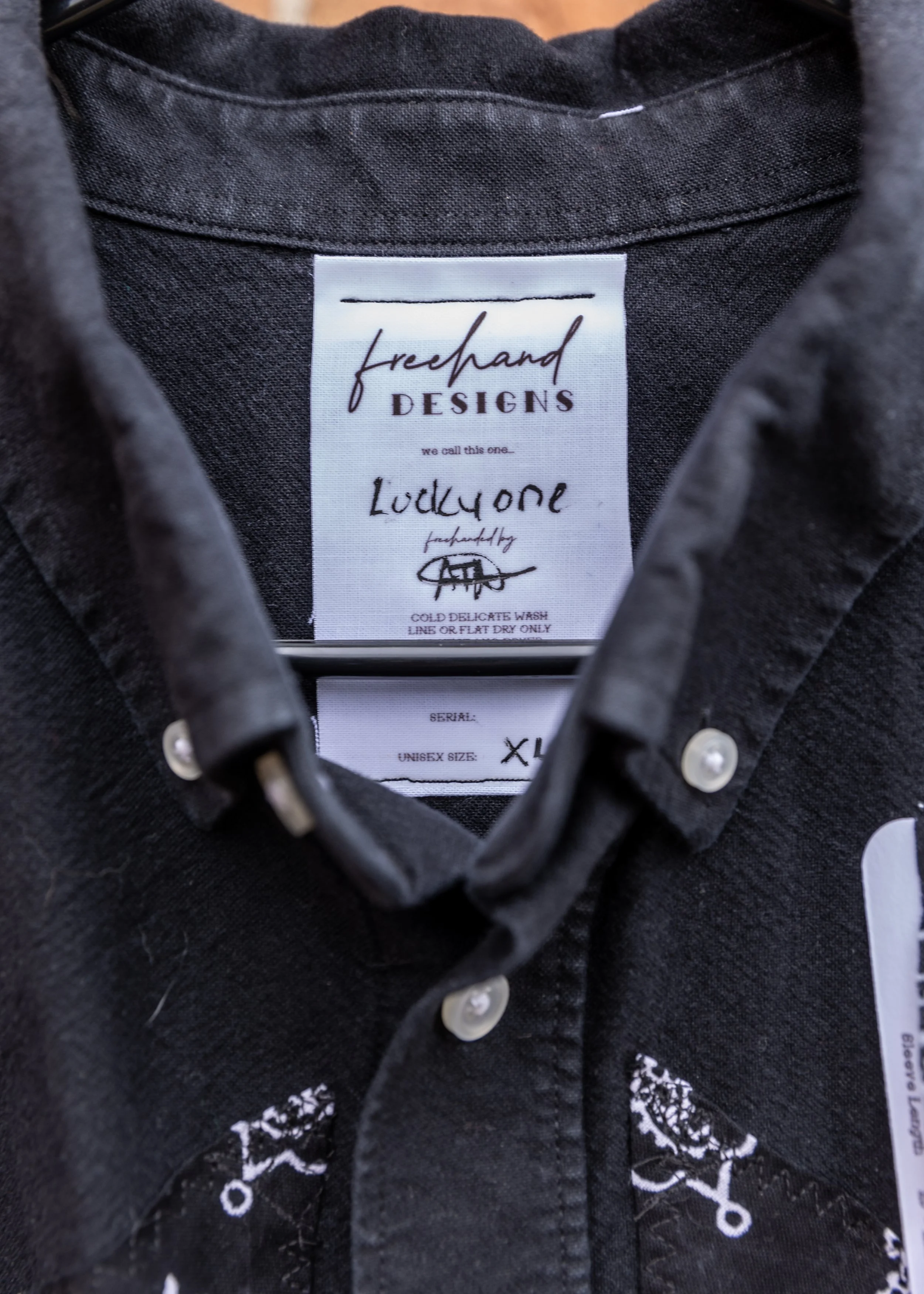 Close-up of a black garment's label that reads 'freehand DESIGNS' and 'Loddyone,' with washing instructions and serial number, surrounded by a black fabric with white embroidery.