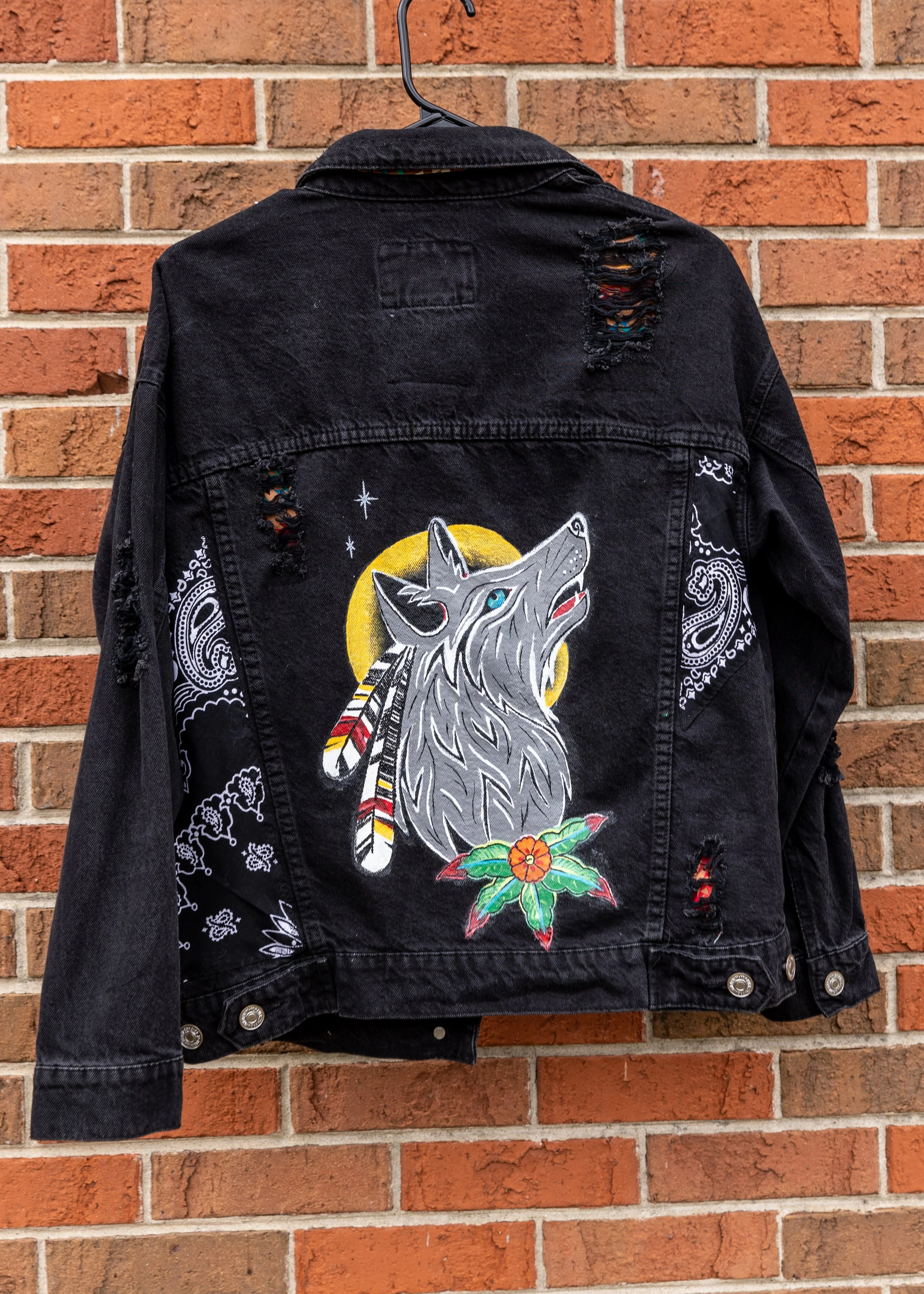 Black denim jacket hangs on a hook against a brick wall. The jacket features a large embroidered wolf head with a moon and feathers, along with intricate white designs on the sleeves and shoulders.
