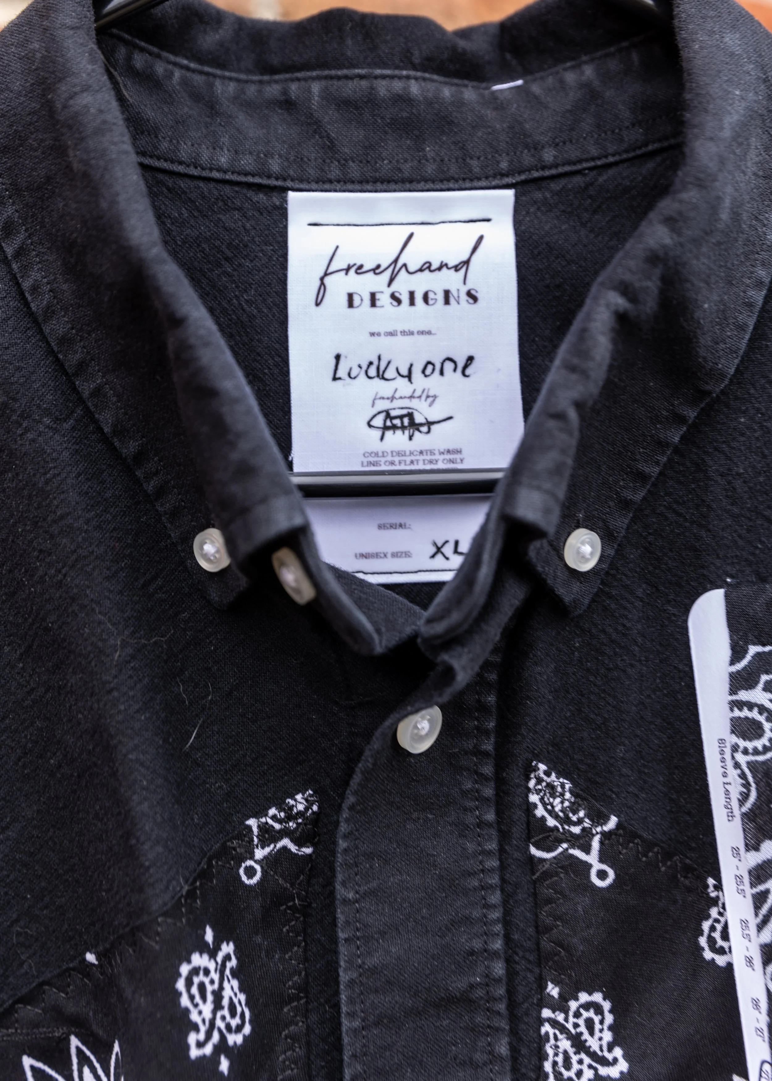 Close-up of black denim shirt with white label inside collar reading 'freehand DESIGNS' and 'Lucky one' with a signature and washing instructions.