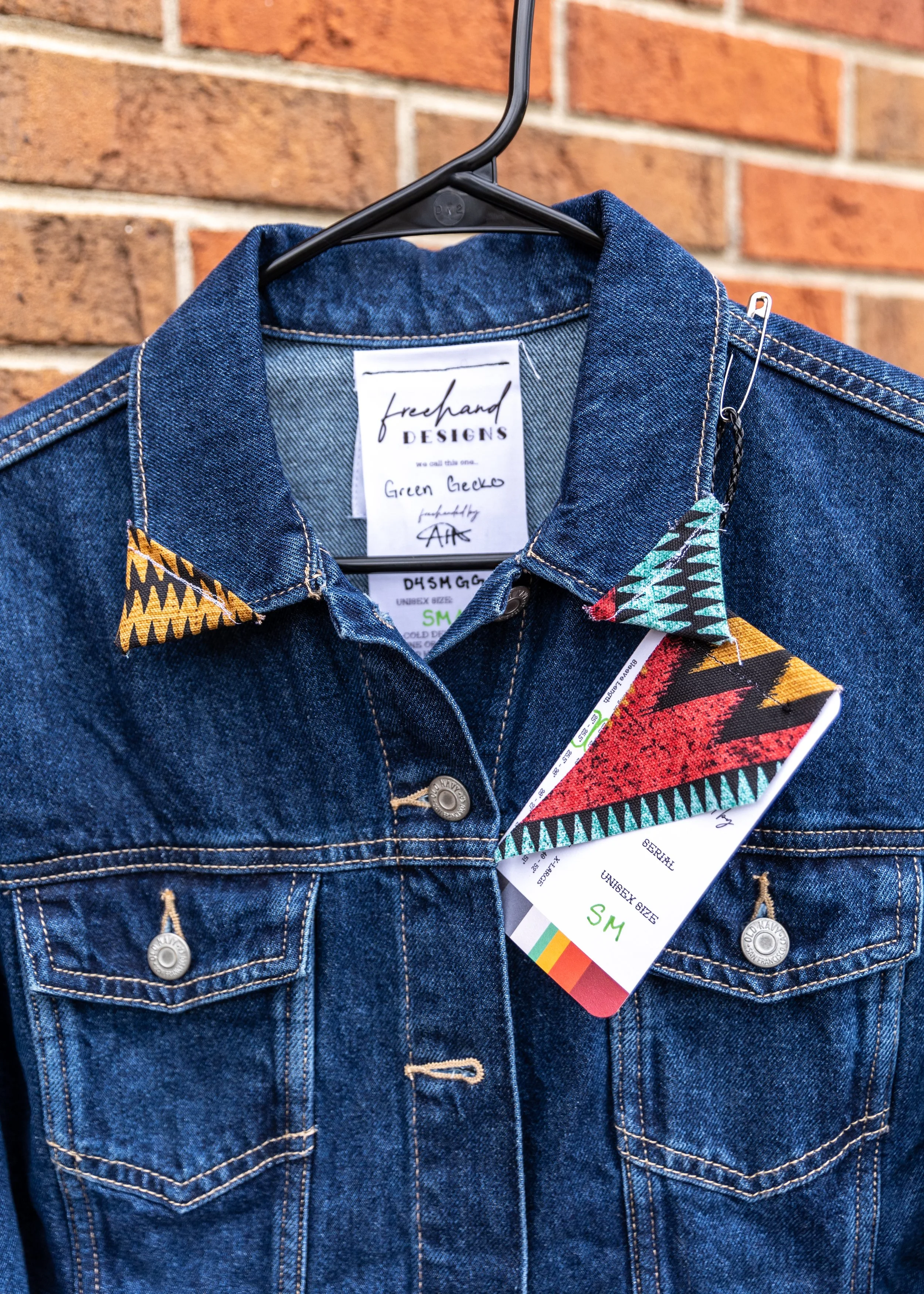 A denim jacket hanging on a black hanger against a brick wall. The jacket has a colorful tag attached and a label that reads 'freehand DESIGNS' with handwritten text 'Green Gecko' and signature.