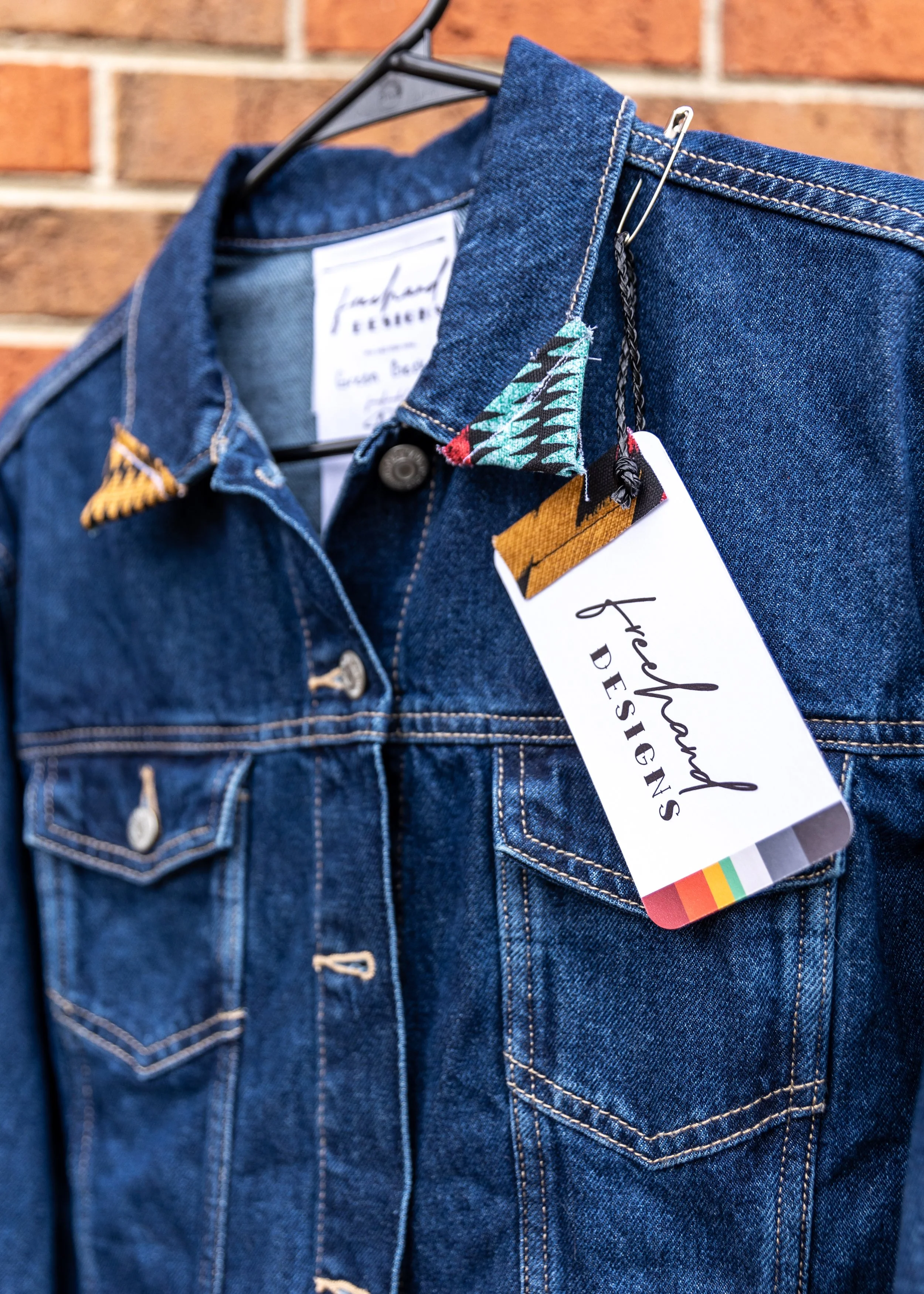 Close-up of a blue denim jacket hanging on a black hanger against a brick wall, with a white tag attached that reads 'Frederick DESIGN' and has a multicolored strip at the bottom.