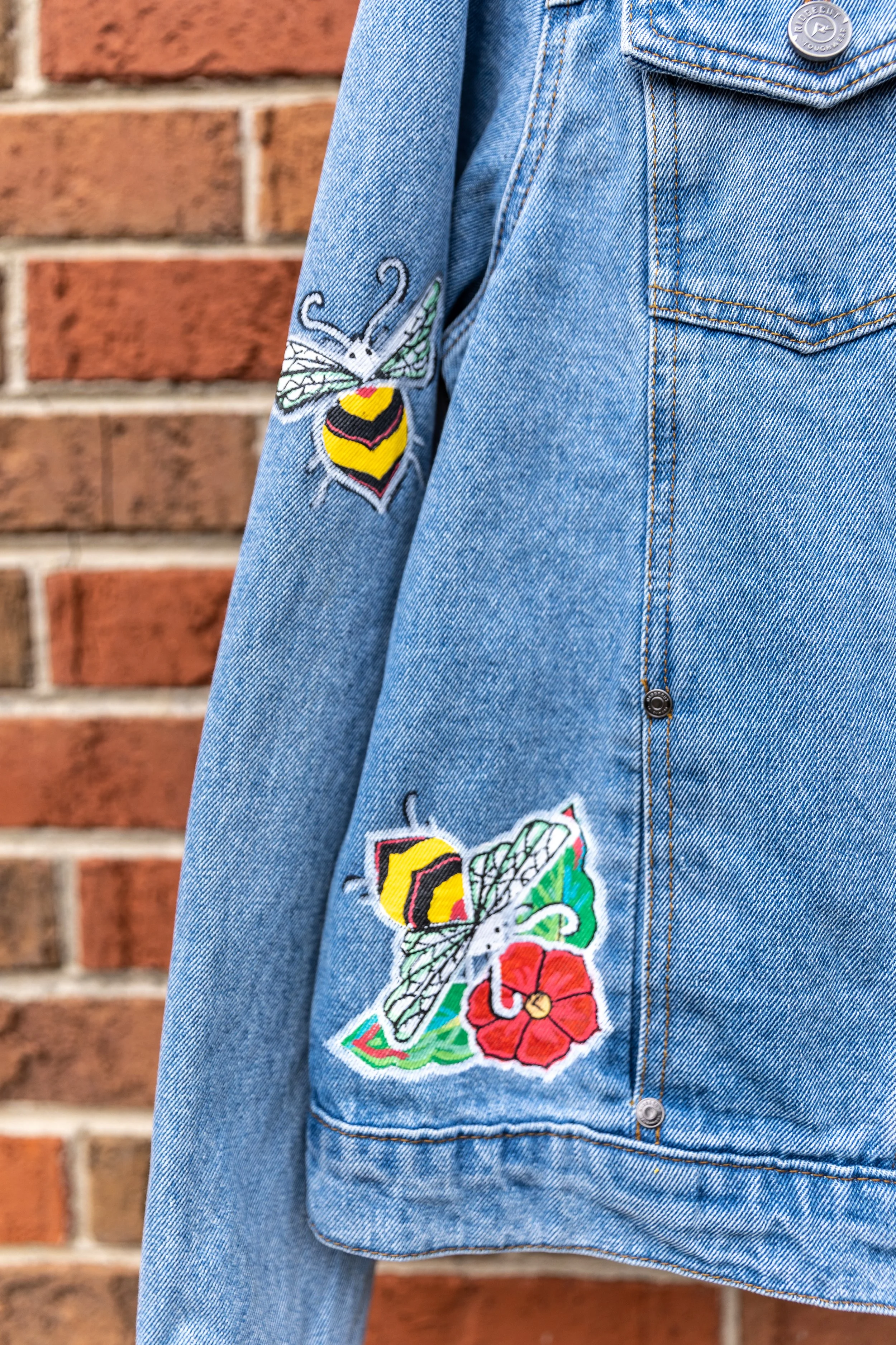 Close-up of a light blue denim jacket with embroidered patches of a bee and a colorful flower design, against a brick wall background.
