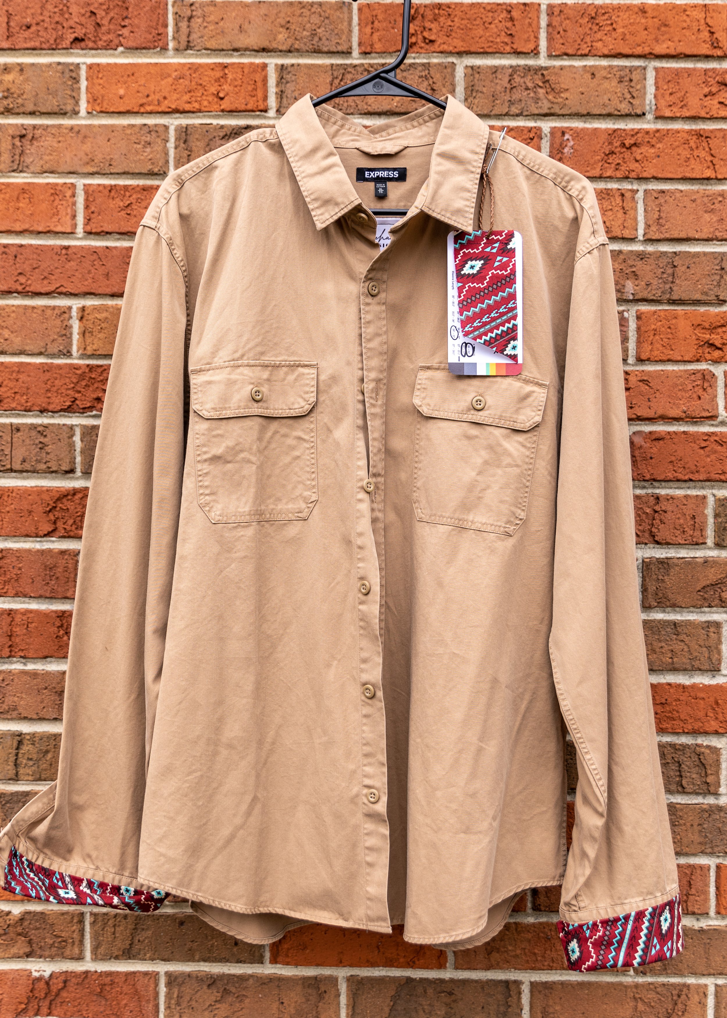 Tan long-sleeve button-up shirt hanging on a black hanger in front of a brick wall, with a colorful band on the inside of the cuffs.