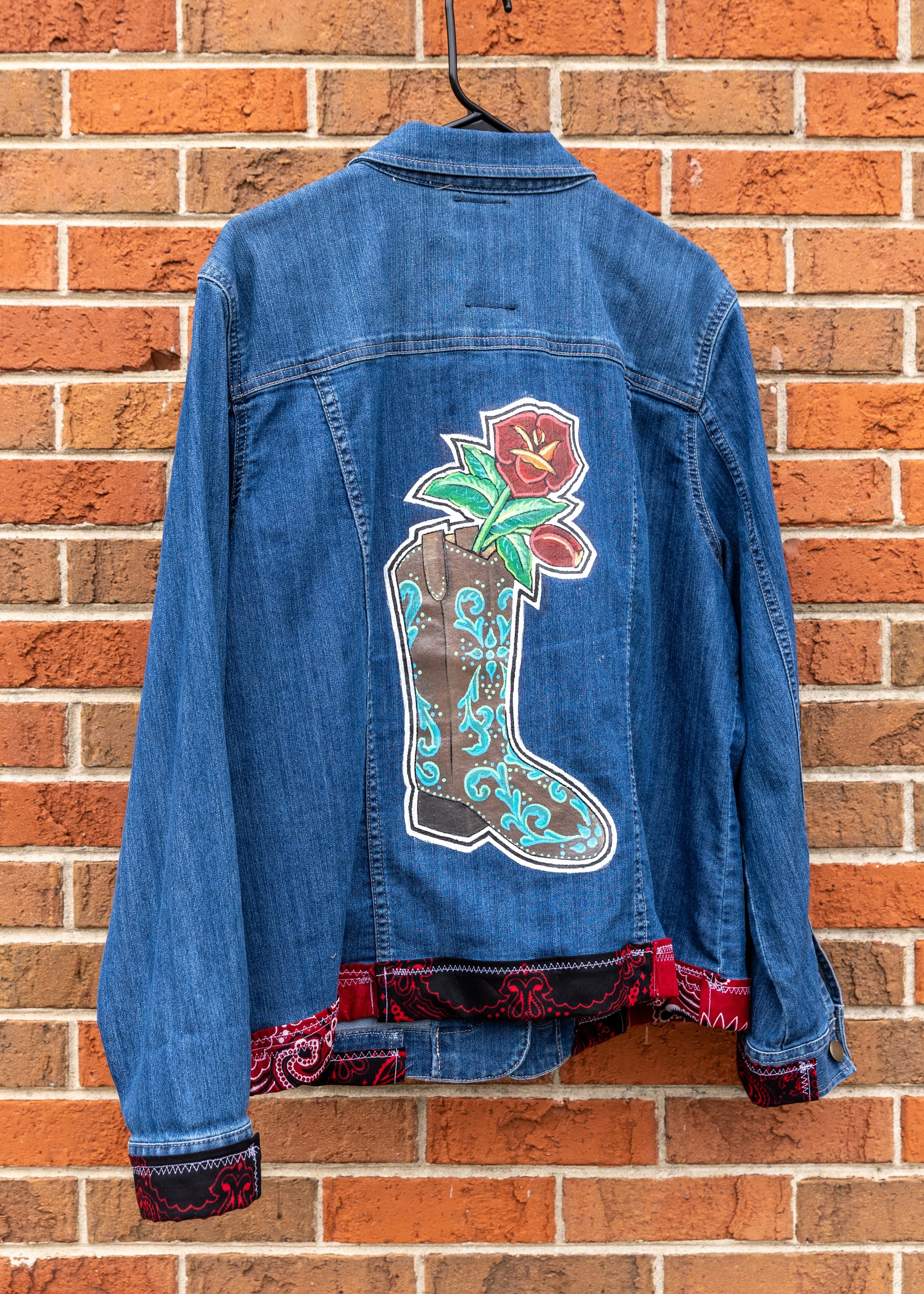 Blue denim jacket with a colorful embroidered design of a flower in a pot on the back, hanging against a brick wall.