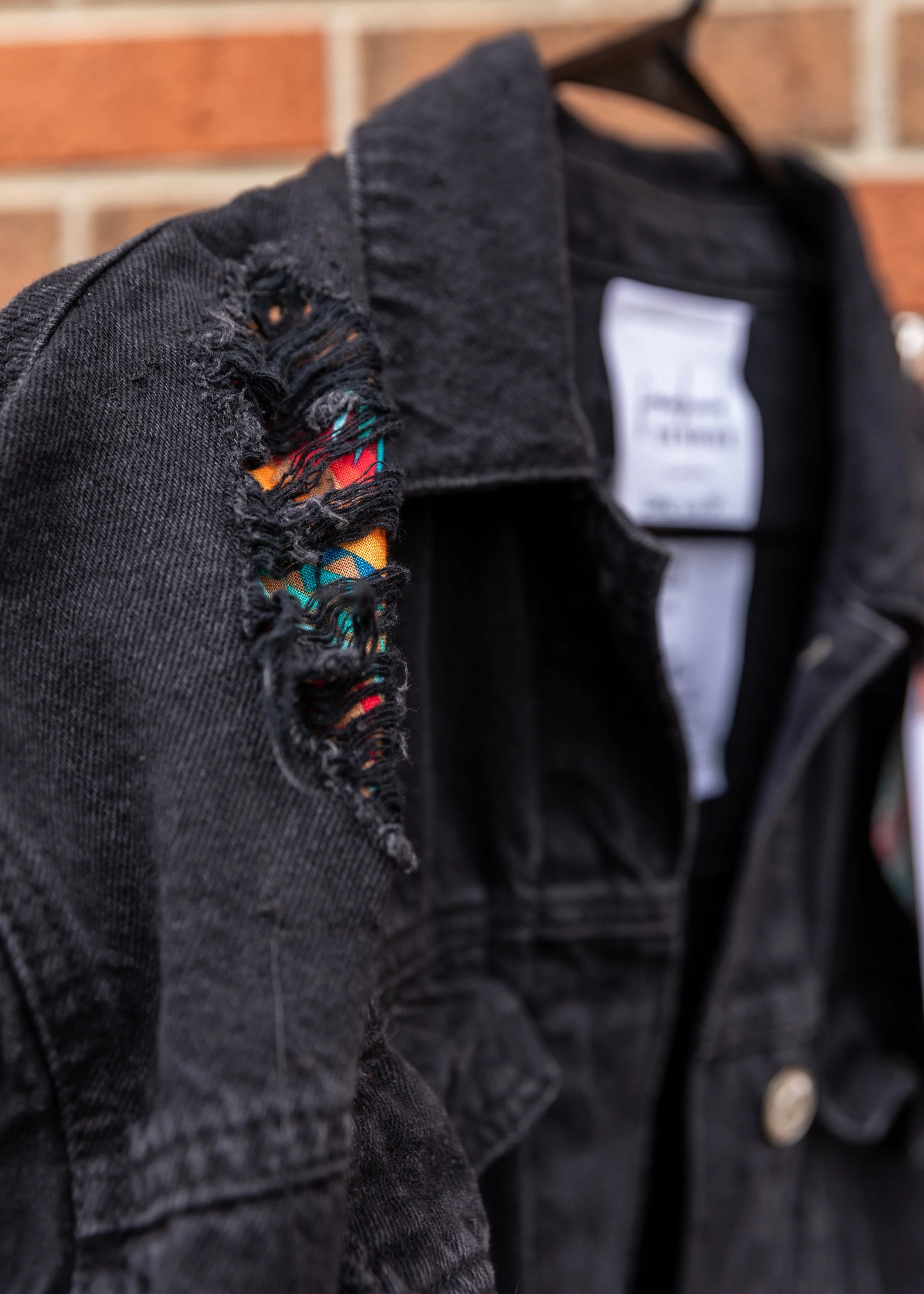 Close-up of a black denim jacket with a distressed torn sleeve, showing colorful fabric underneath, hanging against a brick wall.