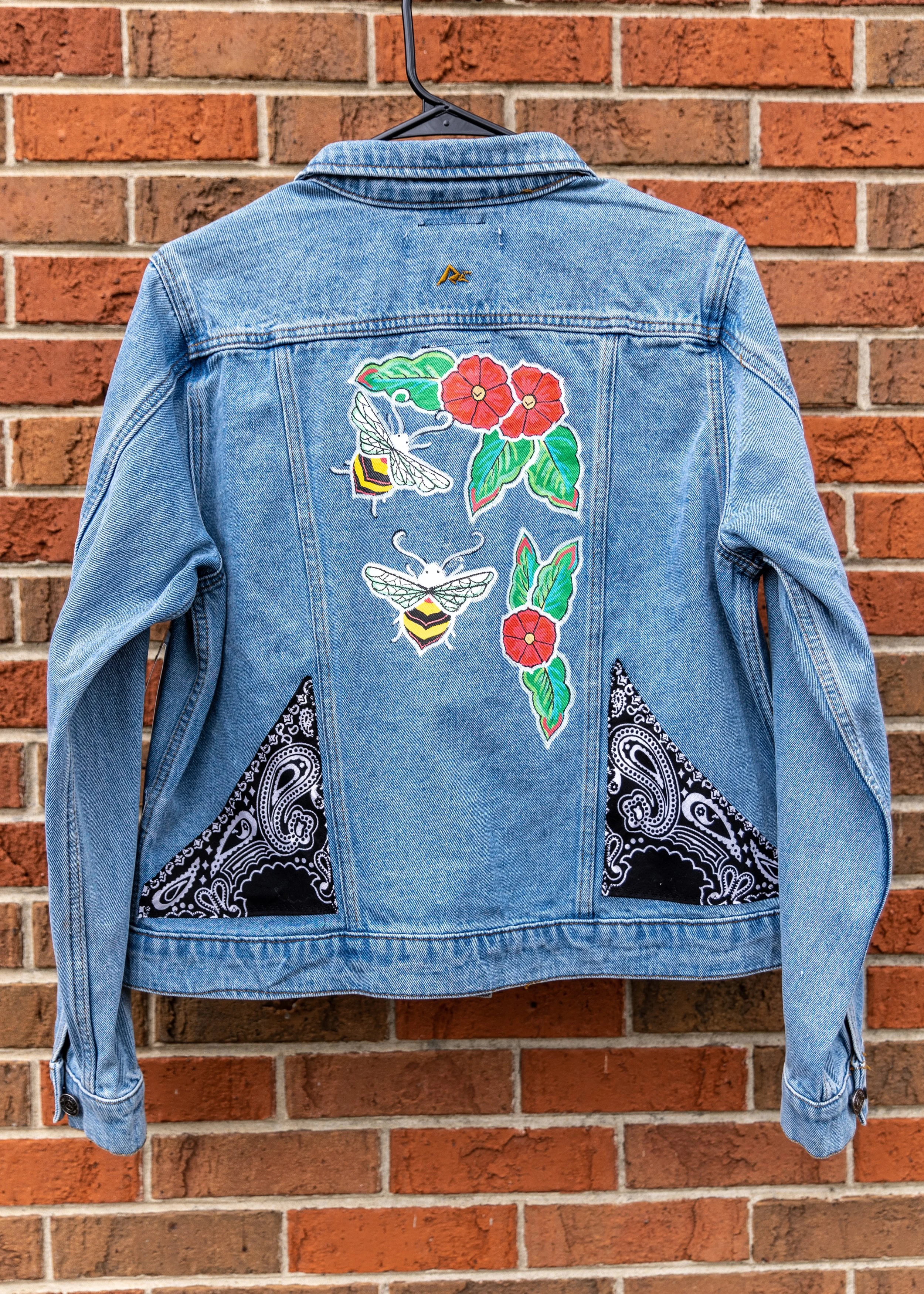 Blue denim jacket with floral and bee embroidery on the back, hanging on a black hanger against a red brick wall.