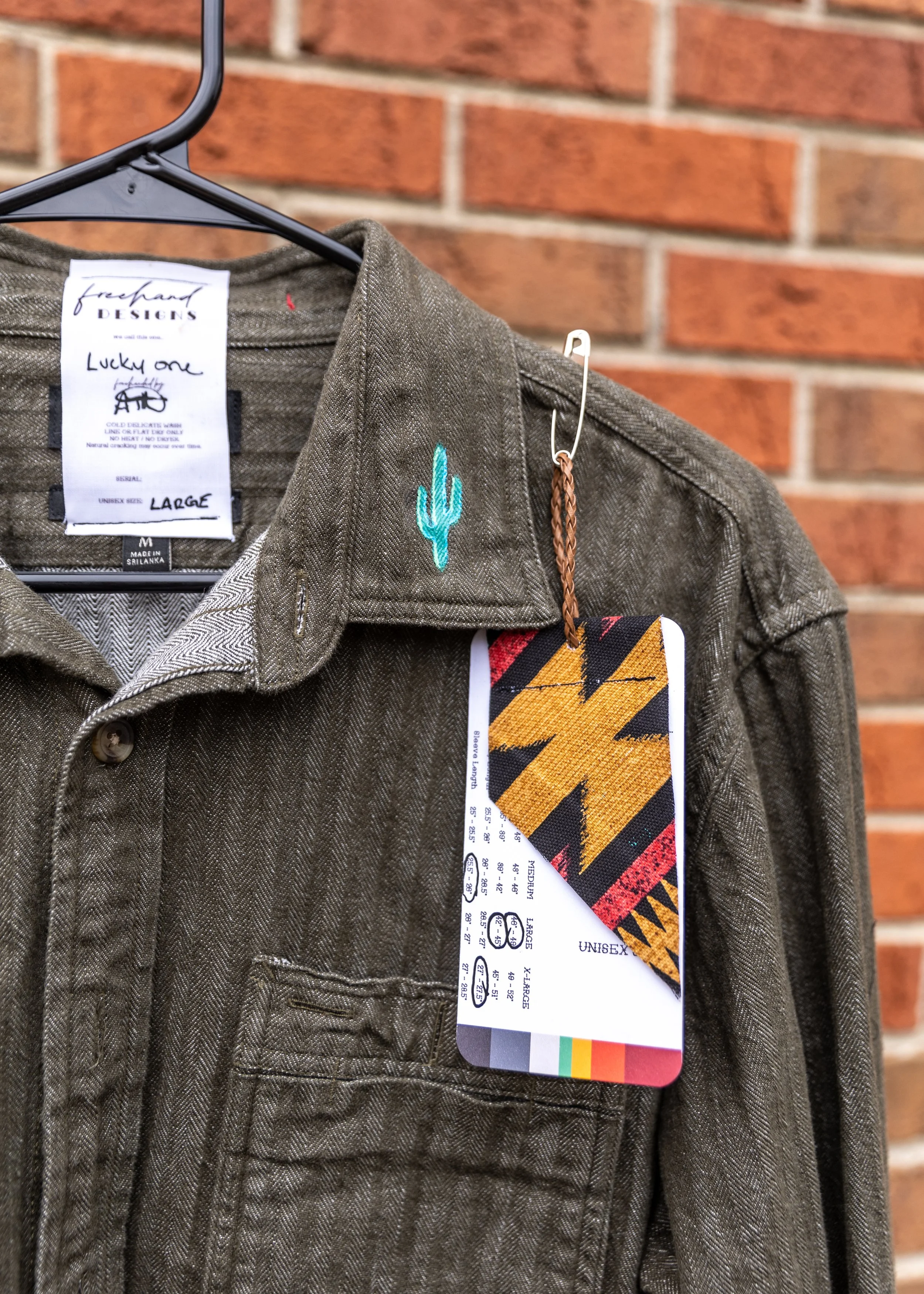A greenish-brown jacket with a cactus embroidery on the shoulder and a colorful tag hanging from it, hanging on a black hanger against a brick wall.