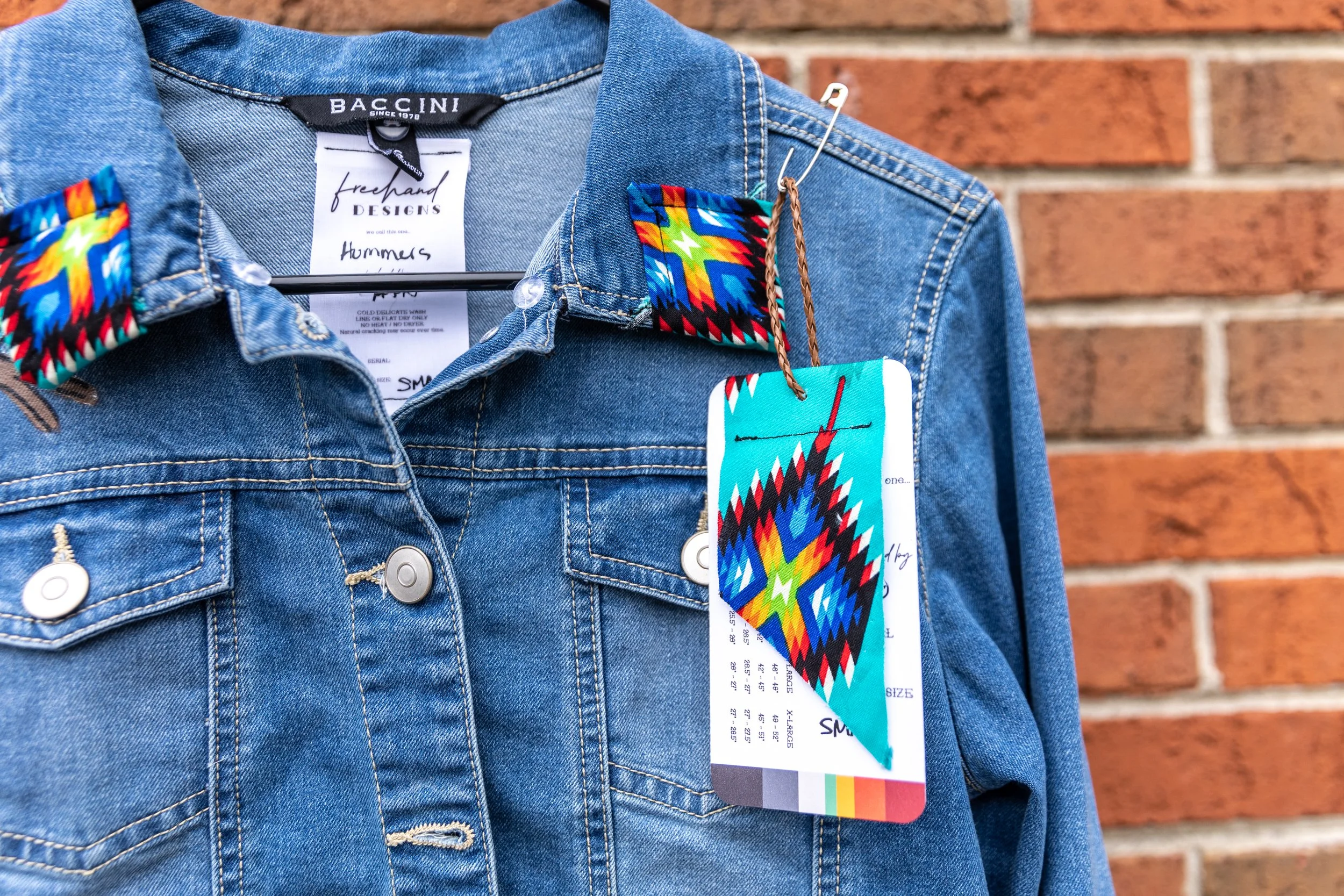 A denim jacket with colorful tribal-patterned accents on the collar and a tag with a geometric design hanging from the pocket against a brick wall.