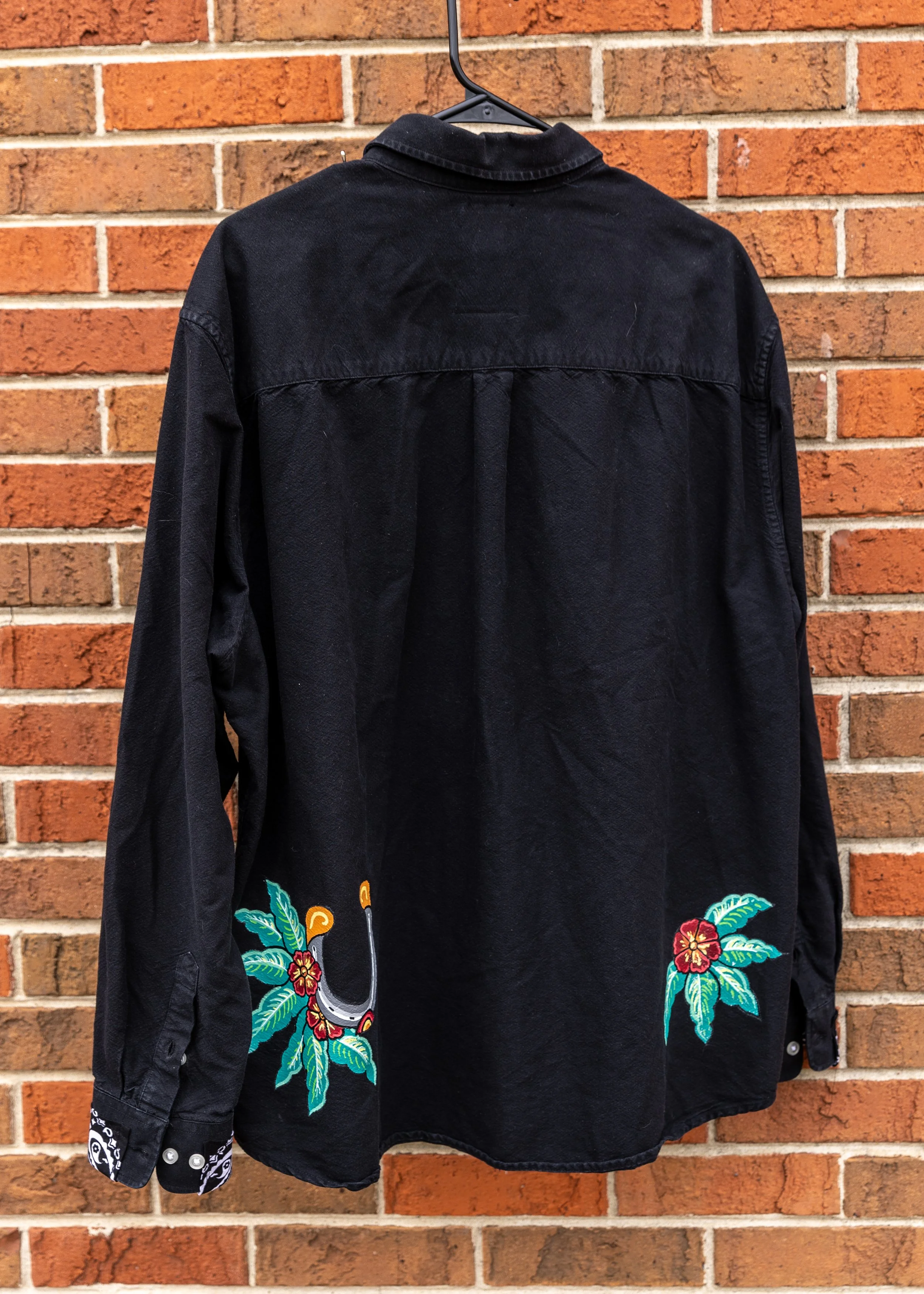 A black denim jacket with colorful embroidery of flowers and a snake on the back, hanging on a hanger in front of a brick wall.