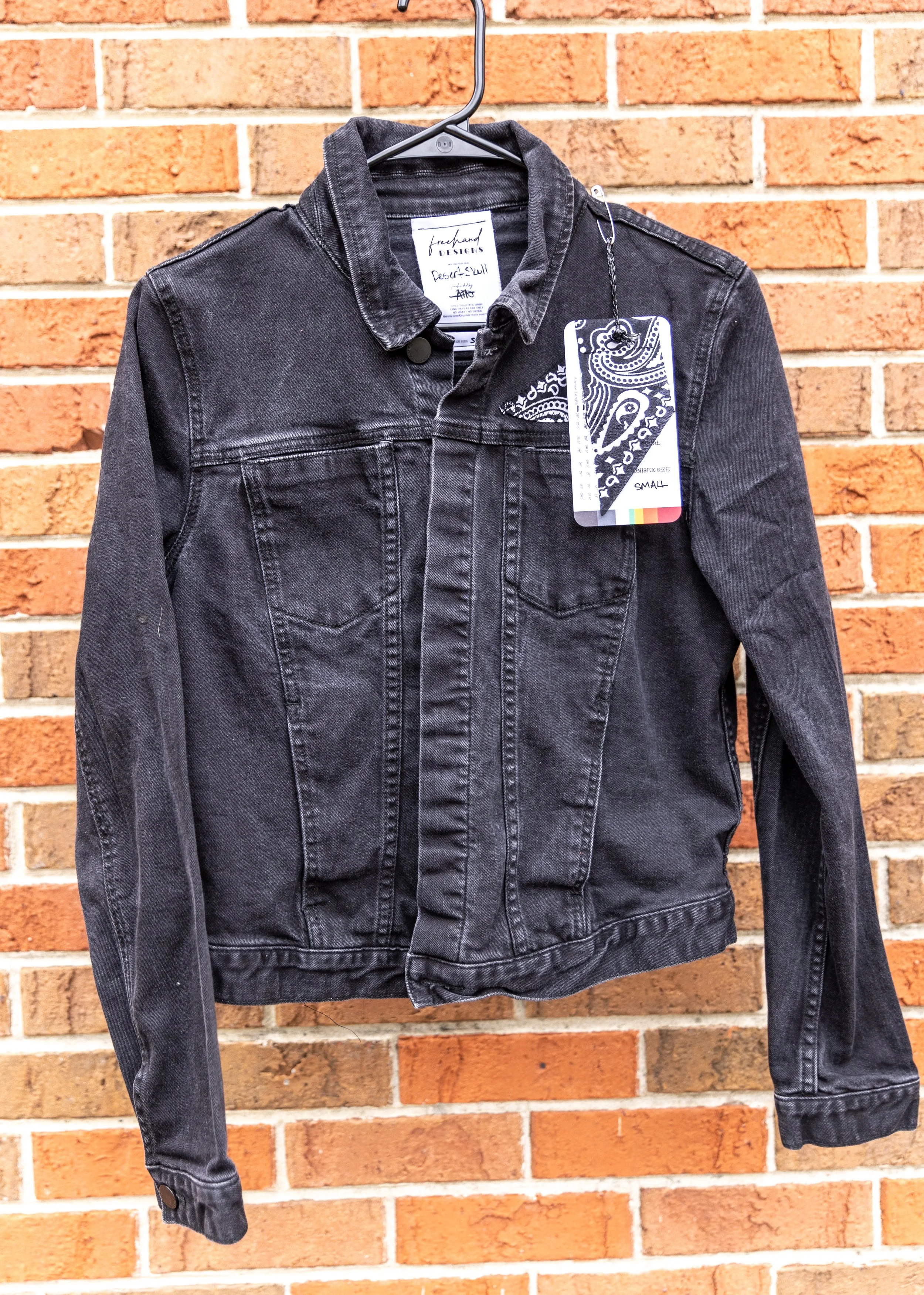 Black denim jacket hanging on a black metal hanger against a brick wall.