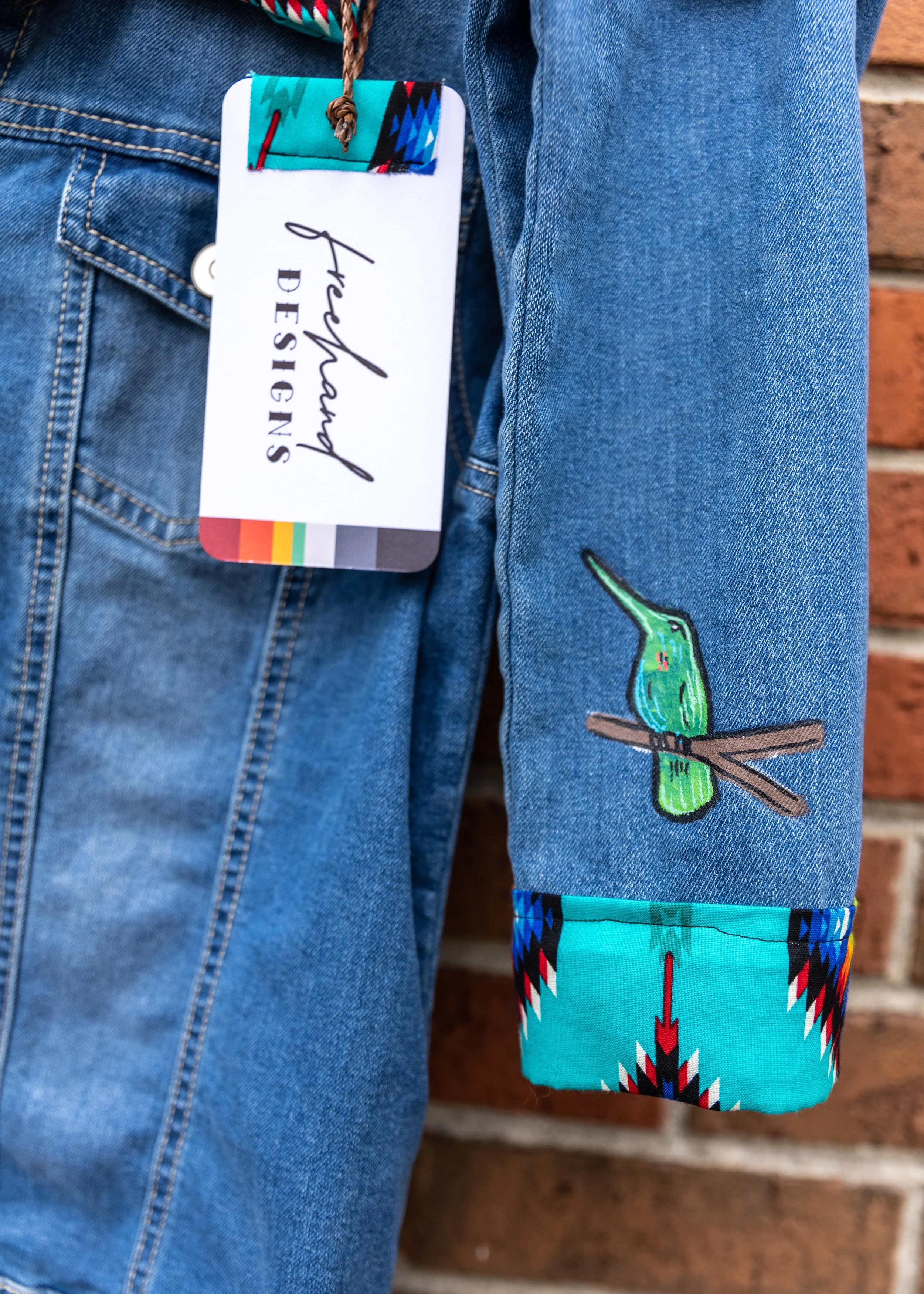 Close-up of a person wearing blue jeans with a colorful tag hanging from the pocket, and a blue denim sleeve with embroidered green dragonfly and geometric patterned fabric on the cuff, in front of a brick wall.