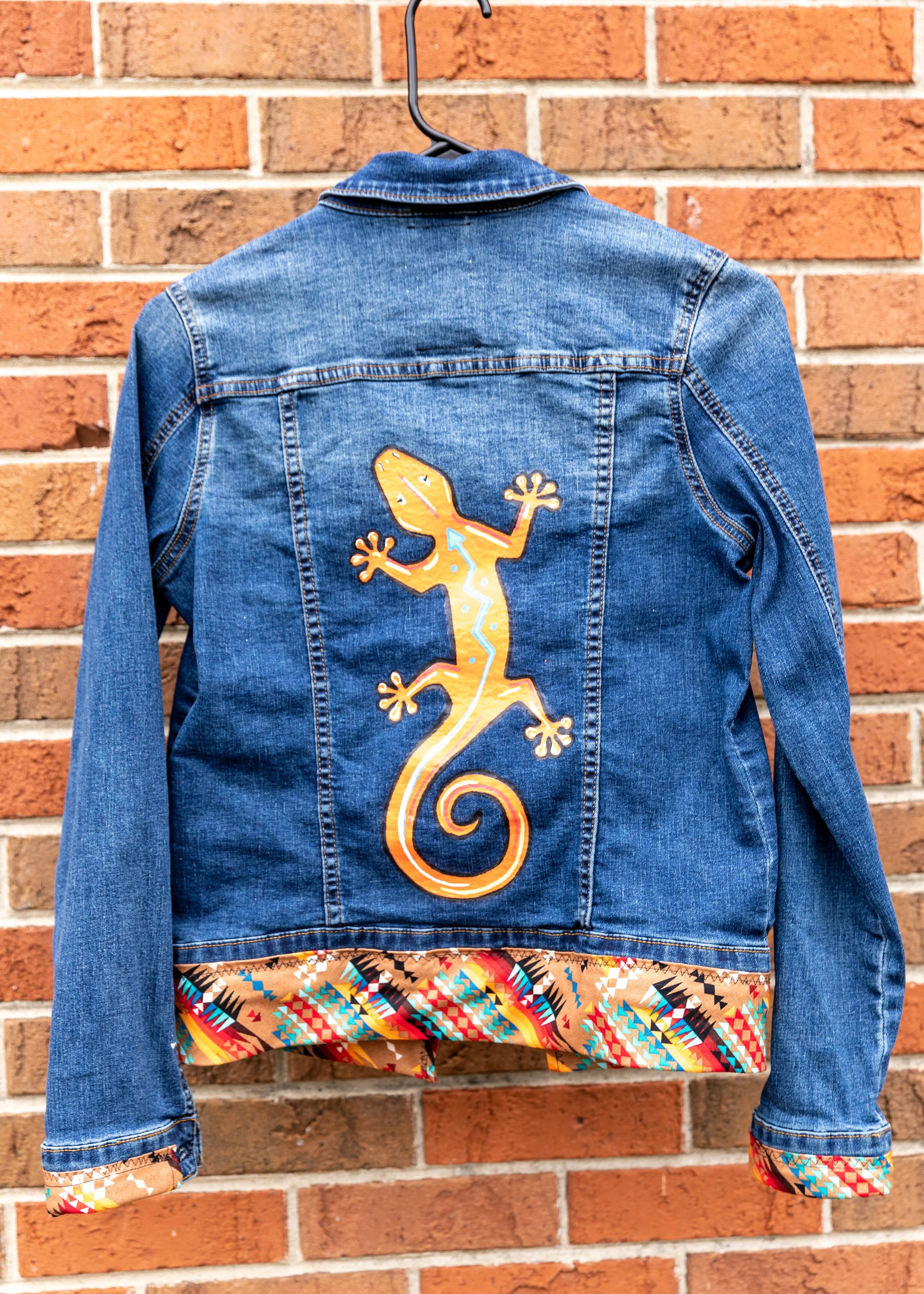 A denim jacket with a colorful gecko patch on the back, hanging on a brick wall.