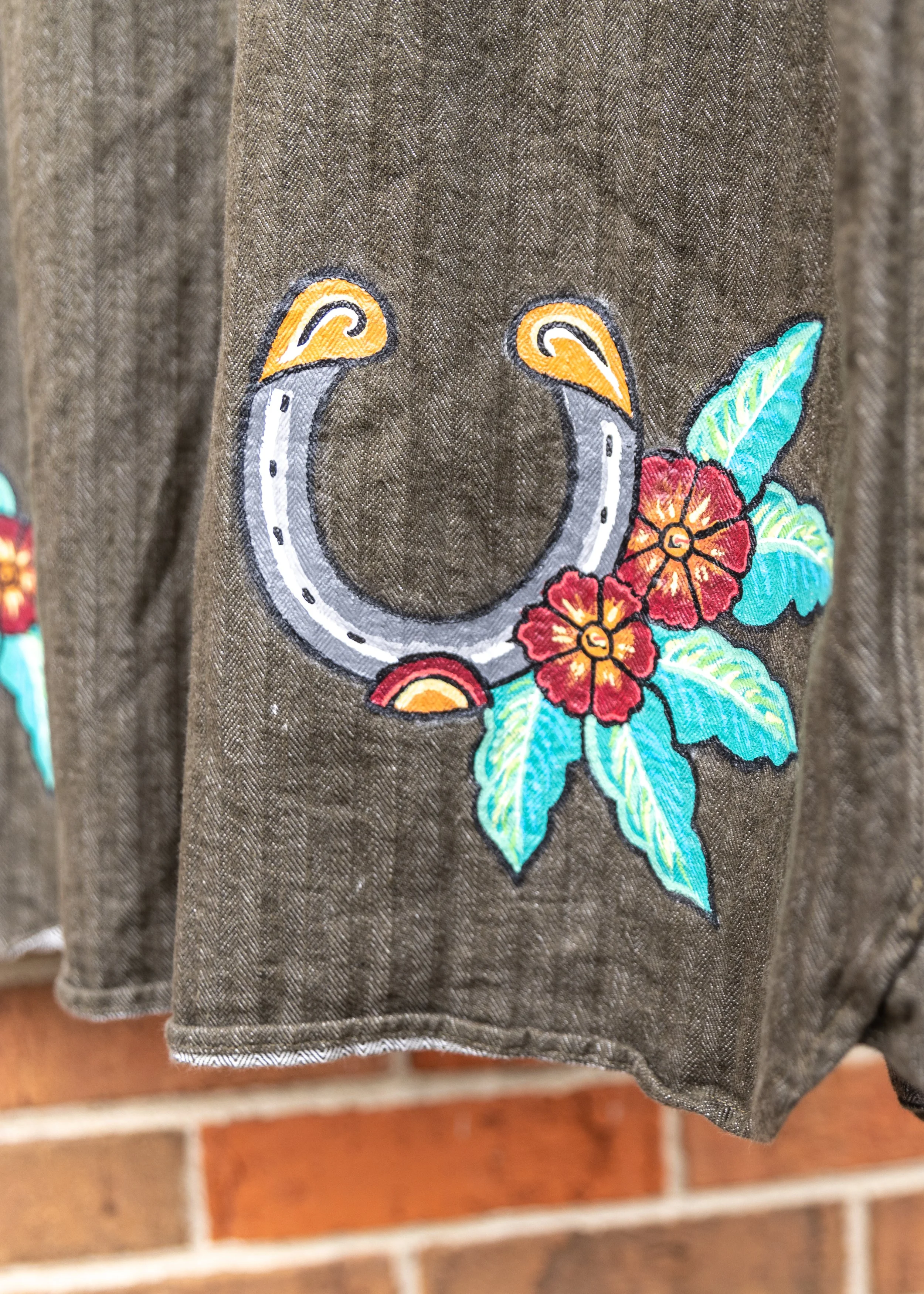 Close-up of embroidered design on brown fabric, featuring a horseshoe, red flowers, and turquoise leaves.