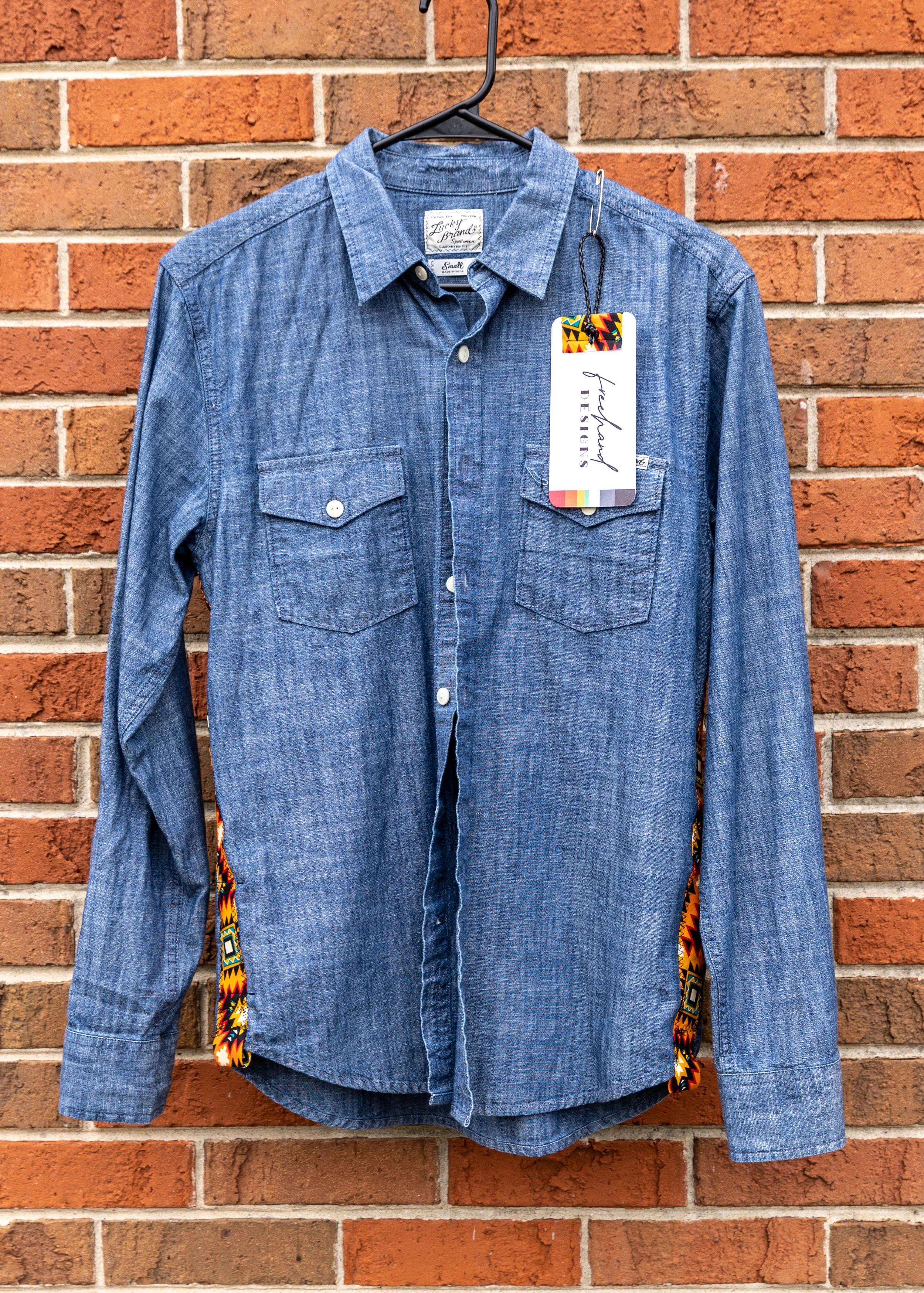 A blue denim casual long-sleeve shirt with two front pockets hanging on a black hanger against a brick wall, with a decorative tag attached.