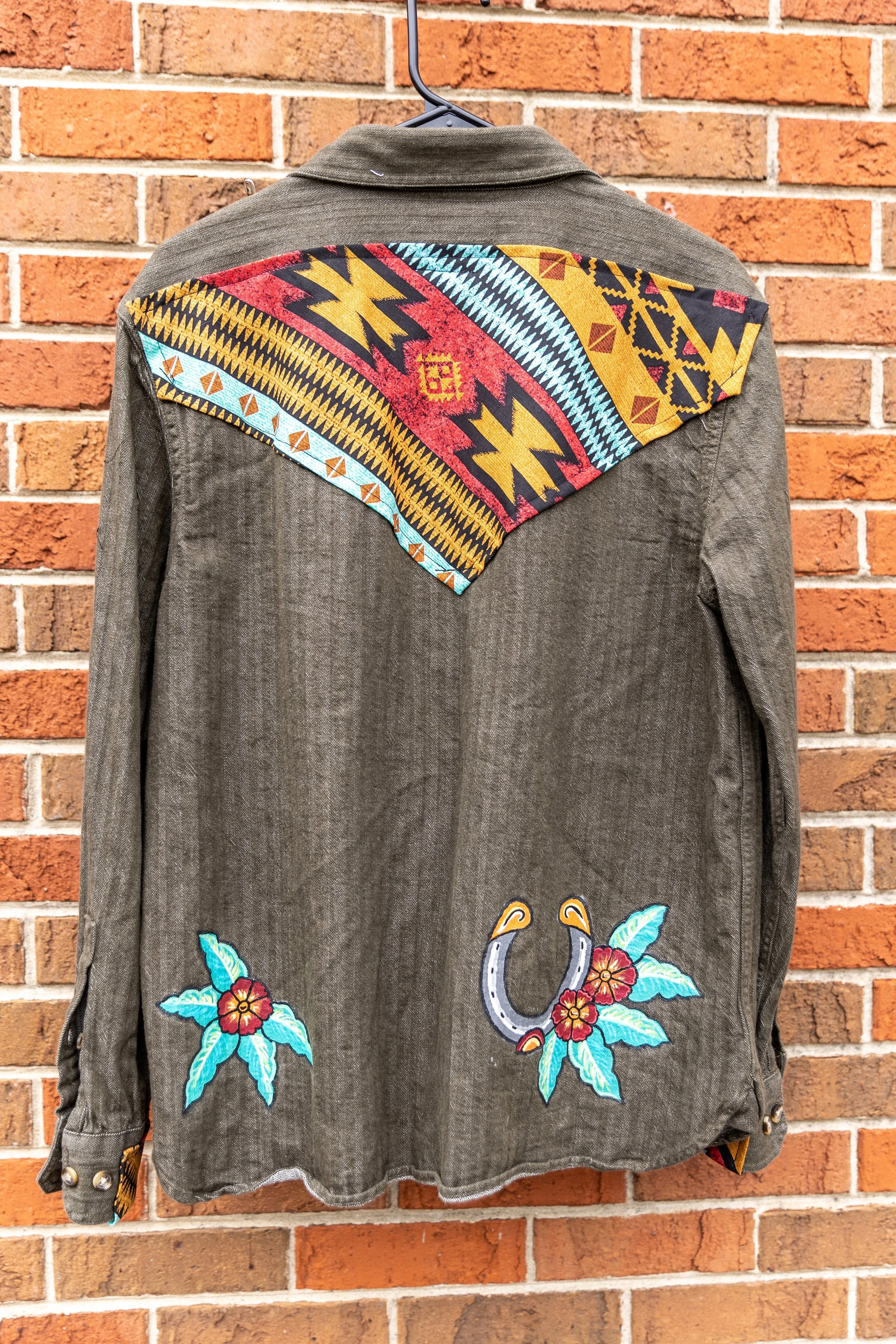 A denim jacket with embroidered colorful floral and animal designs hanging on a hanger against a brick wall.