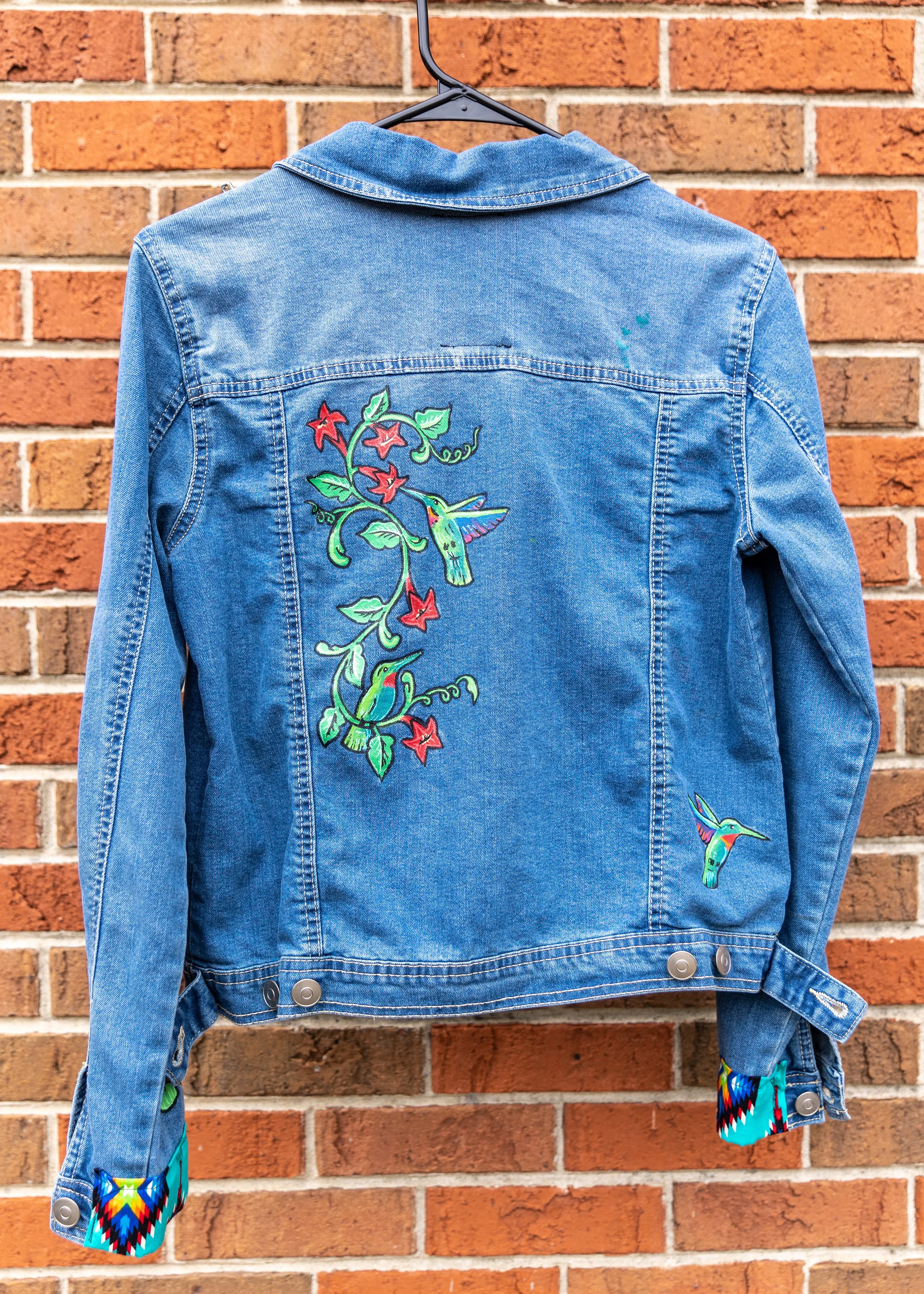A blue denim jacket with colorful embroidery of a hummingbird and flowers hanging on a black hanger against a brick wall.