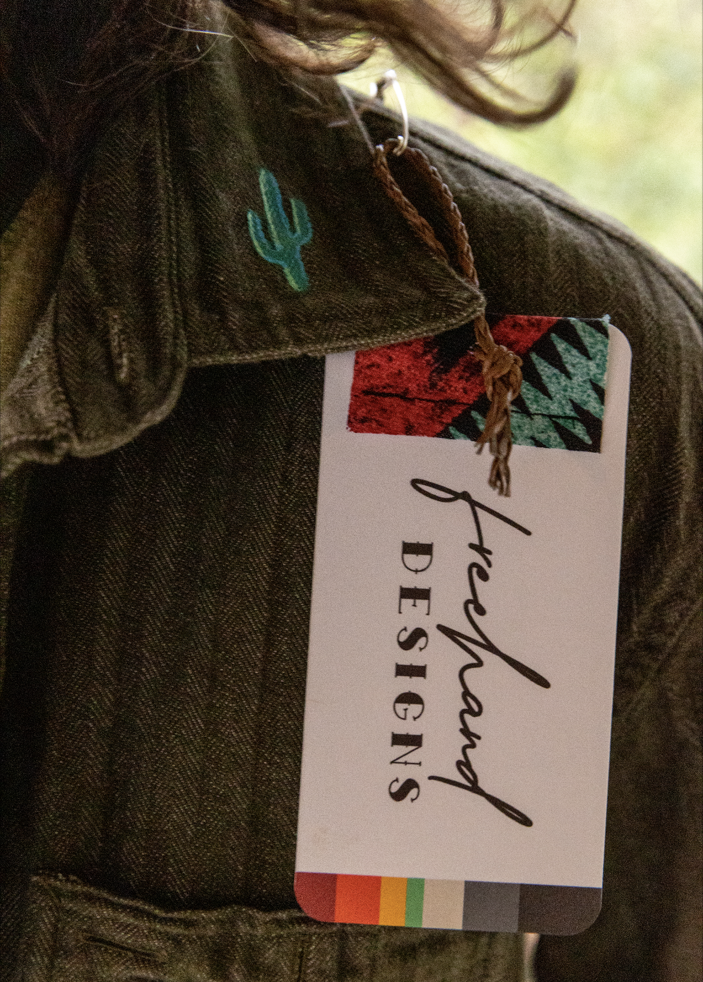 Close-up of a biohazard hazard tag attached to a dark brown leather bag with a cactus embroidered patch, and a colorful striped ribbon at the bottom.