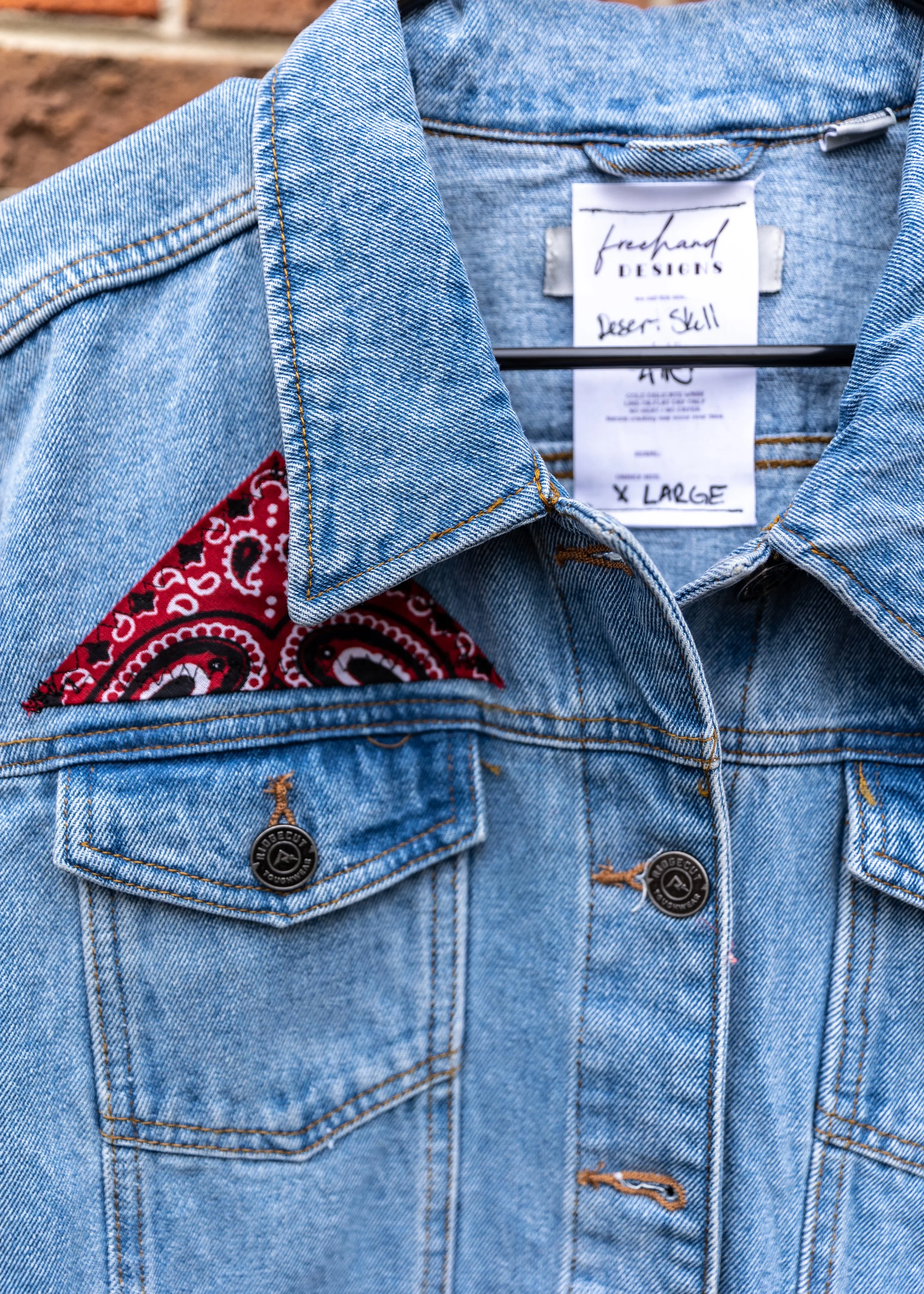 Close-up of a light blue denim jacket showing the collar, pocket, and part of a red bandana pattern inside the pocket. The jacket has metal buttons and a brand tag that reads 'freehand DESIGNS'. The label inside the jacket indicates a size of extra l