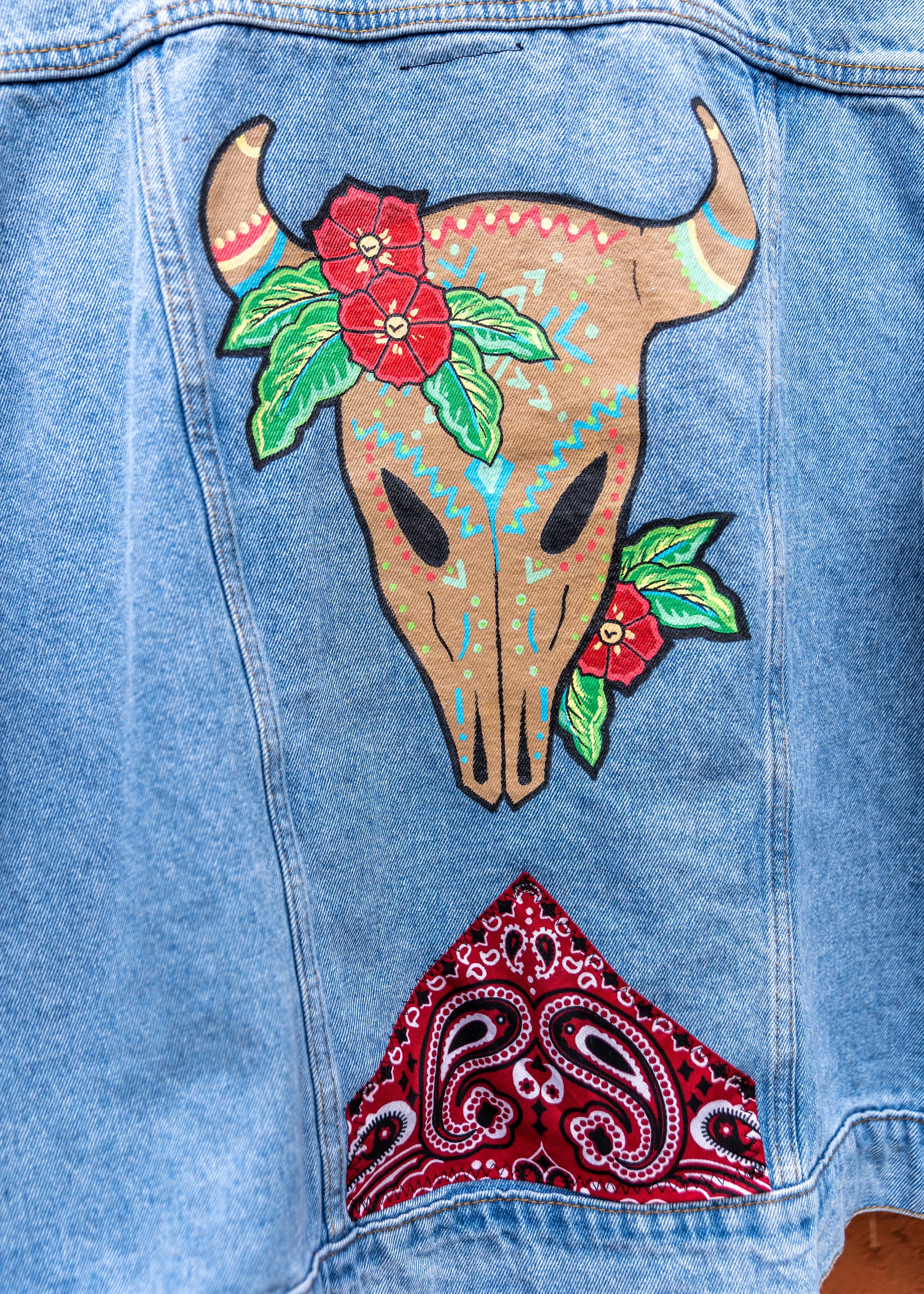 Close-up of a denim jeans back with painted designs resembling a bull skull with red flowers and green leaves, and a red bandana pattern on a pocket.