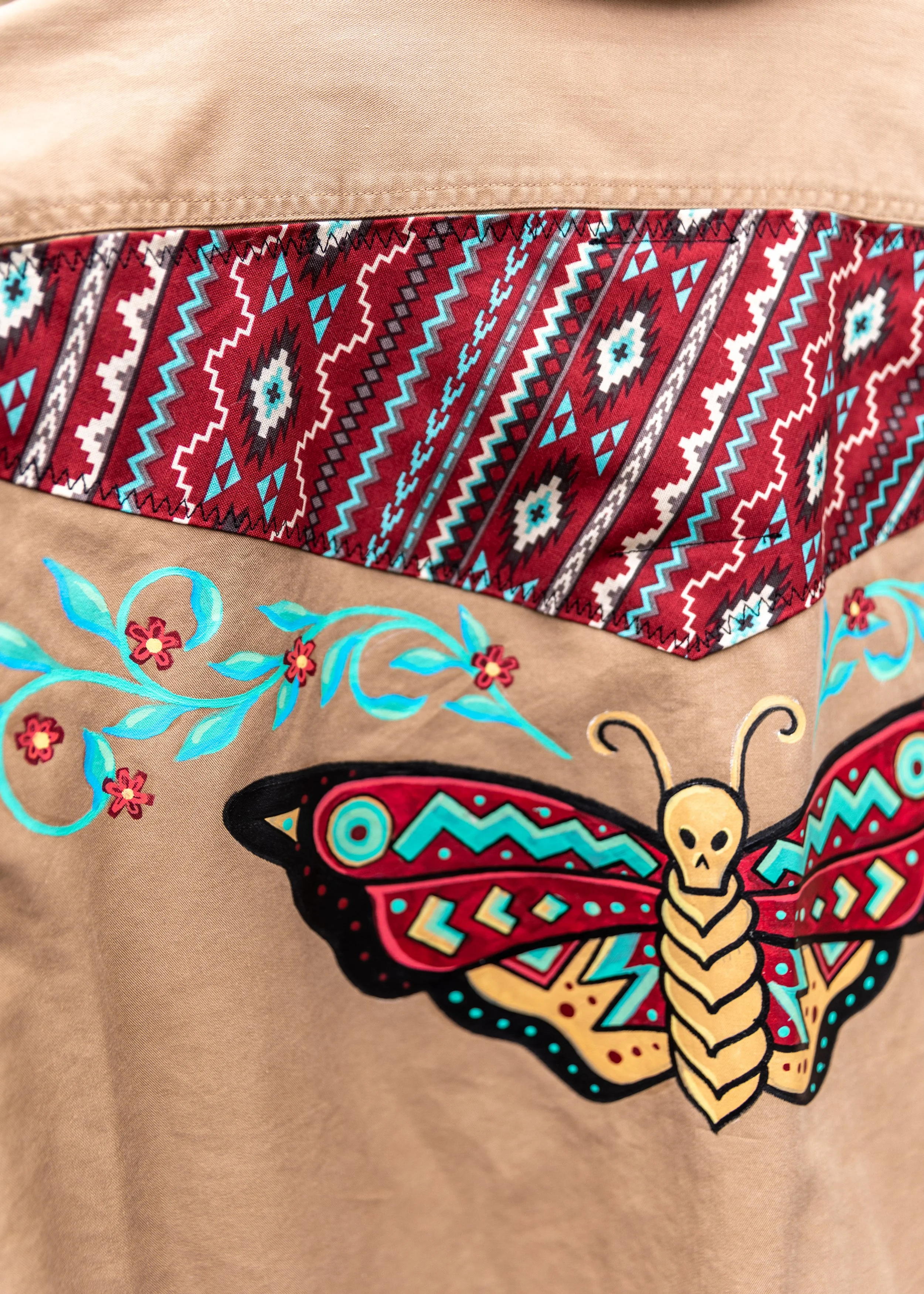 Close-up of fabric featuring a colorful embroidered butterfly with floral and geometric patterns, and a section of woven textile with intricate tribal designs in red, black, white, and blue, set on a tan background.