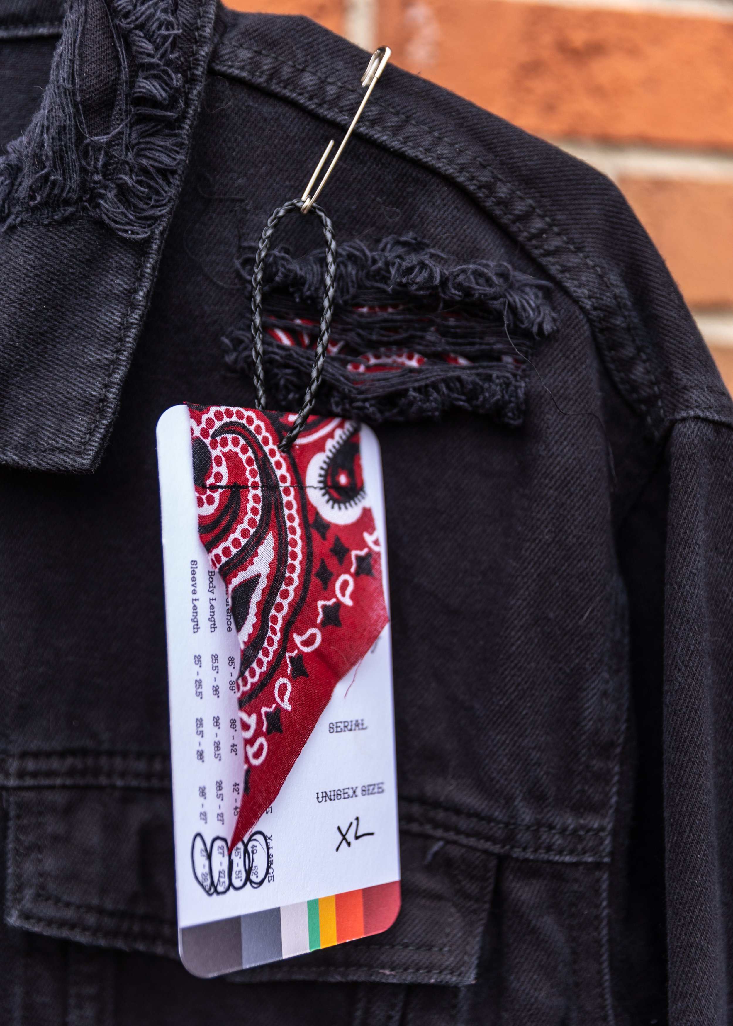 A black denim jacket with a torn distressed style, featuring frayed edges and a tag hanging from the pocket.
