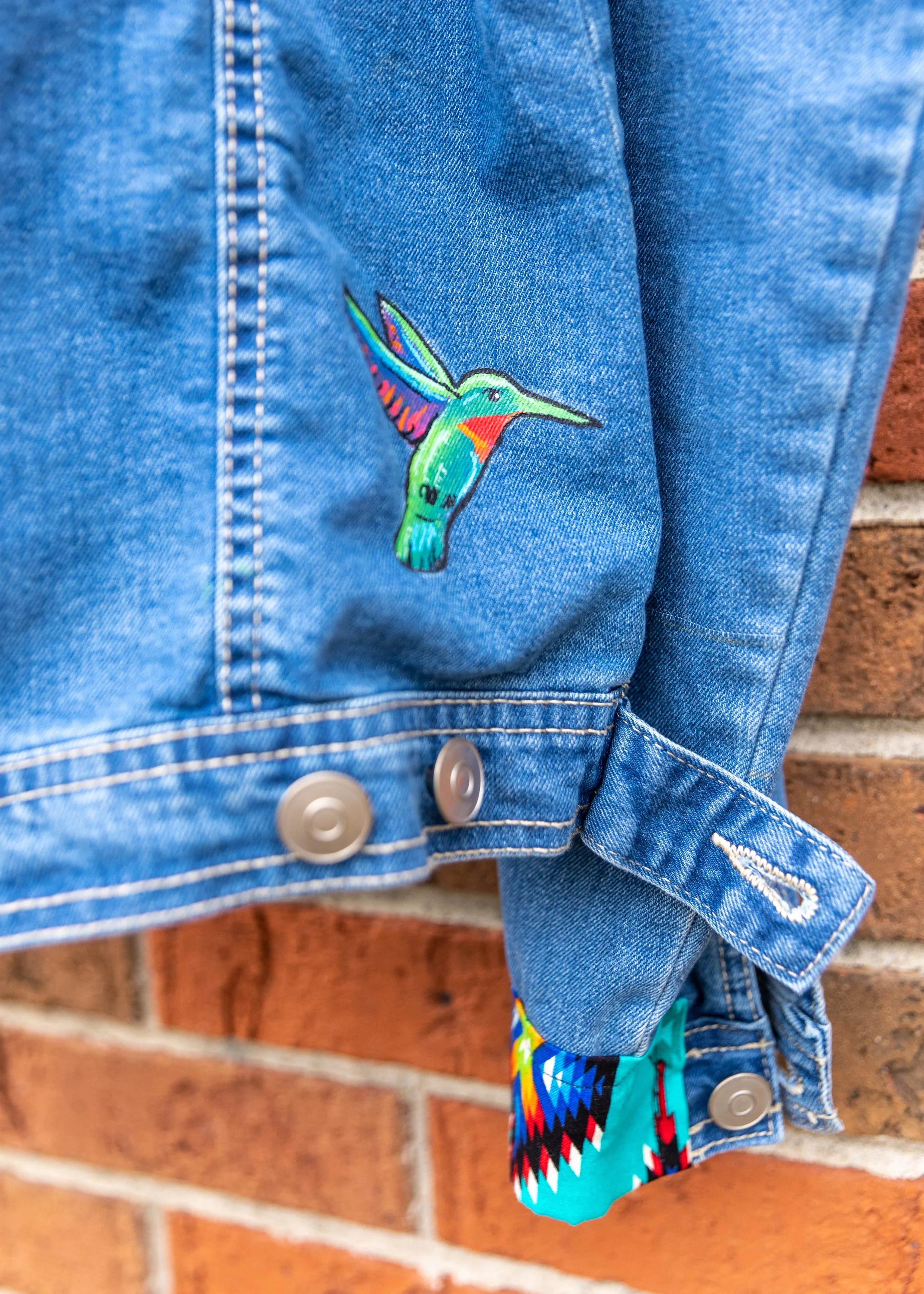 Close-up of a denim jacket with a colorful embroidered hummingbird patch. The jacket is buttoned, and part of a multicolored patterned fabric is visible at the sleeve cuff. The background features a red brick wall.