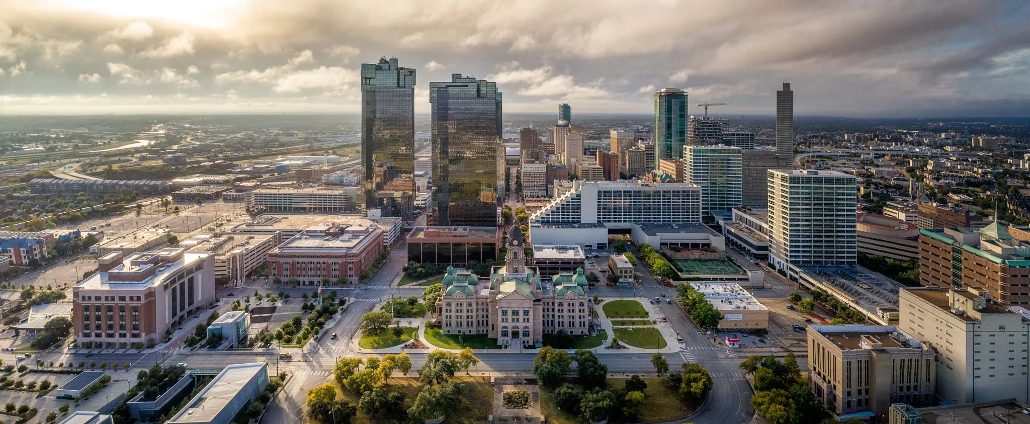 Aerial view of downtown city skyline at sunset featuring modern high-rise buildings, green parks, and a historic-style government building with a green dome in the foreground.