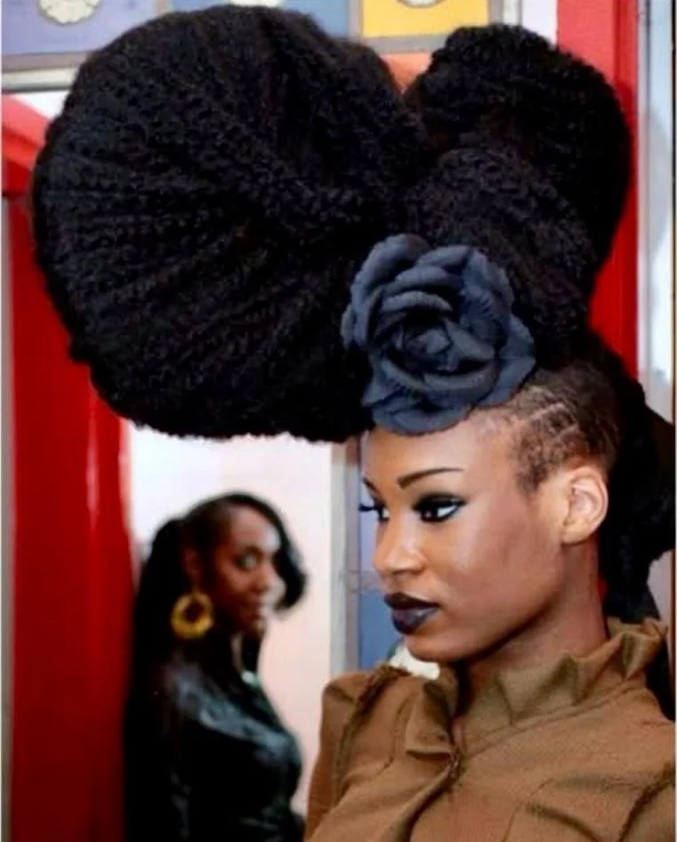 A woman with dark lipstick and eye makeup wearing a brown top with a high collar, and a large elaborate hairstyle with thick black twists arranged in a bun, adorned with a fabric flower, standing inside a bus or train with another woman blurred in th