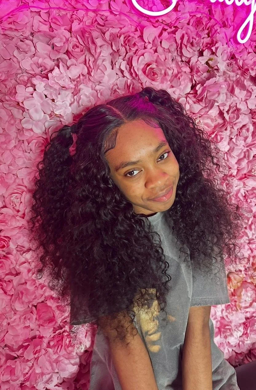 A young woman with curly black hair styled in two pigtails with purple highlights, smiling slightly, sitting against a pink floral wall background.