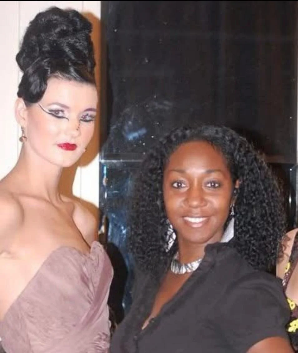 Two women posing indoors, one with elaborate hairstyle and makeup wearing a strapless dress, and the other with curly hair, smiling wearing earrings and a necklace.