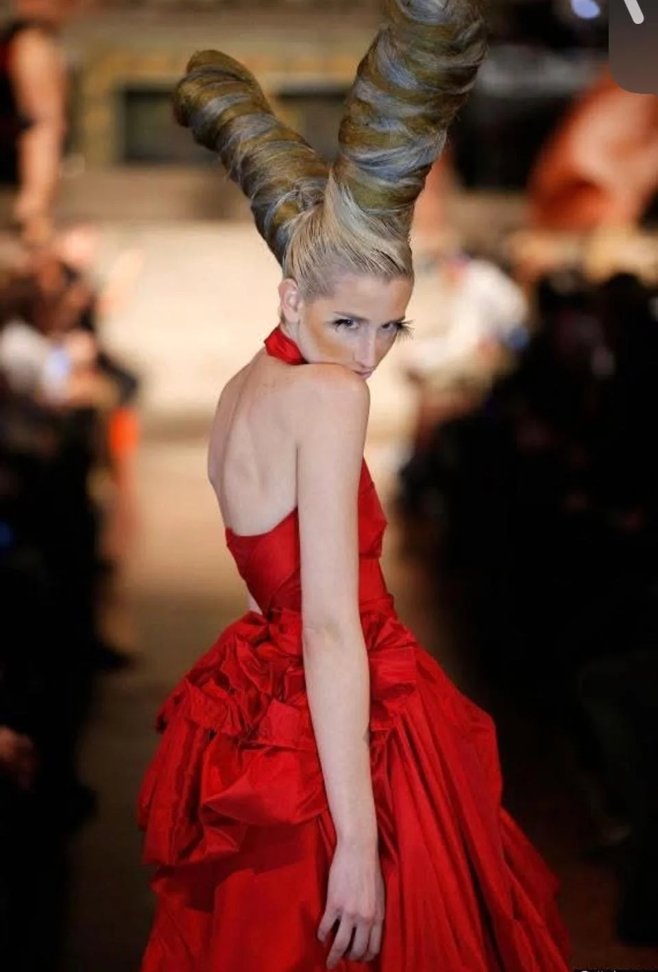 A woman walking on a fashion runway with elaborate twin-updo hairstyle, wearing a red gown with ruffled details, in front of a blurred audience.