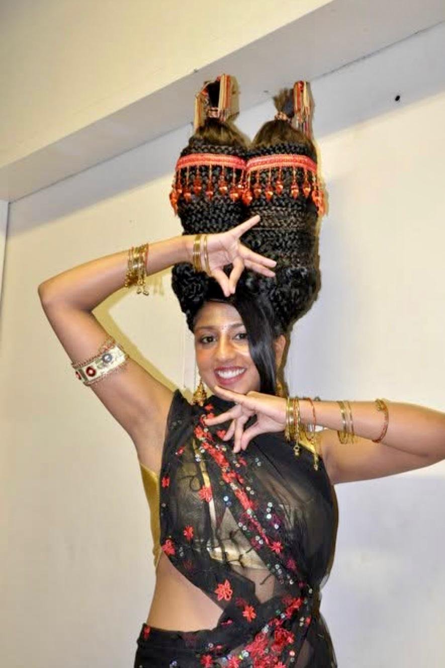 Woman dressed in traditional Indian attire, performing a dance pose with her arms and hands in intricate positions, smiling at the camera.