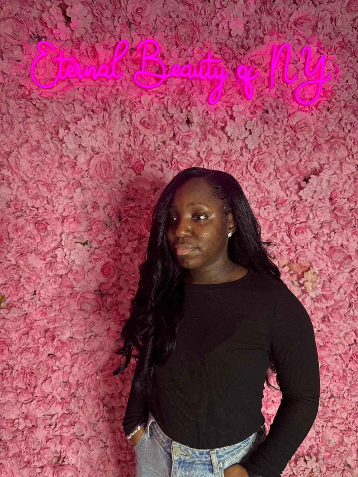 A woman with long black hair, wearing a black top and light blue jeans, stands in front of a pink flower wall with a neon sign that reads 'Eternal Beauty of NY'.