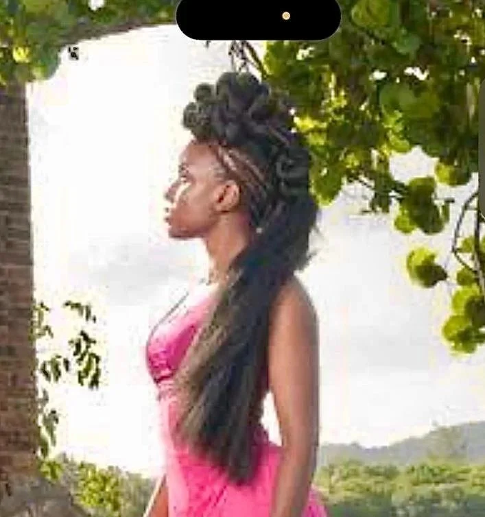 A woman with long braided hair styled with bantu knots, standing outdoors under a tree with green leaves, wearing a pink dress, facing left.